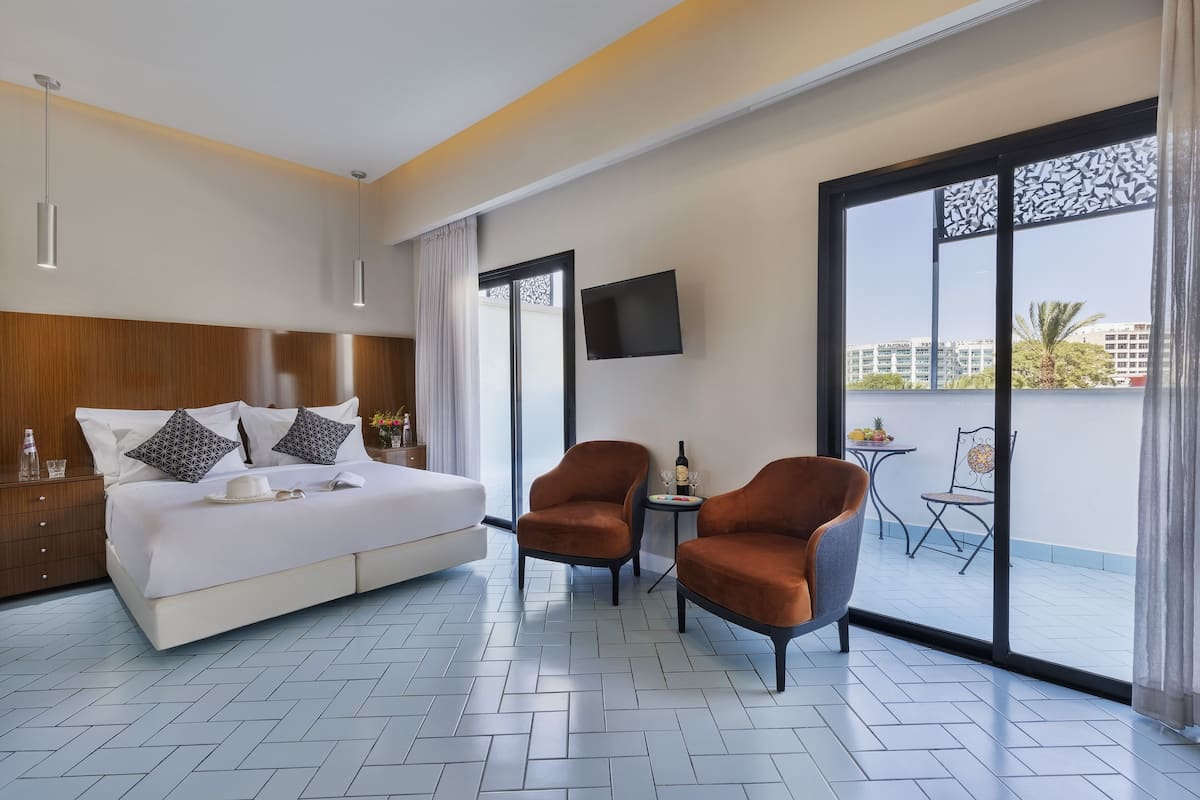 Une chambre d’hôtel moderne avec un grand lit, deux fauteuils, une petite table avec une bouteille et des verres, et un téléviseur fixé au mur.