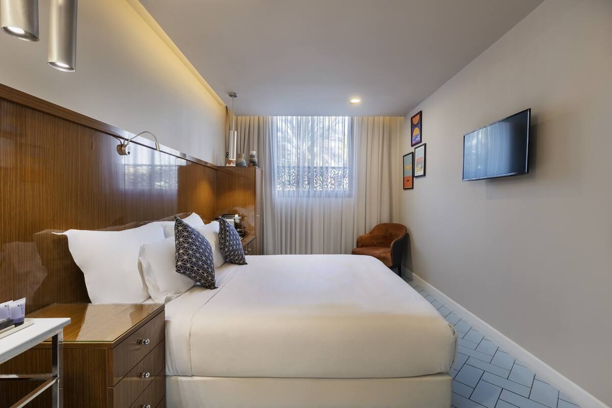 Une chambre d’hôtel avec un lit, une table de chevet, une lampe, une télévision et une fenêtre avec des rideaux.