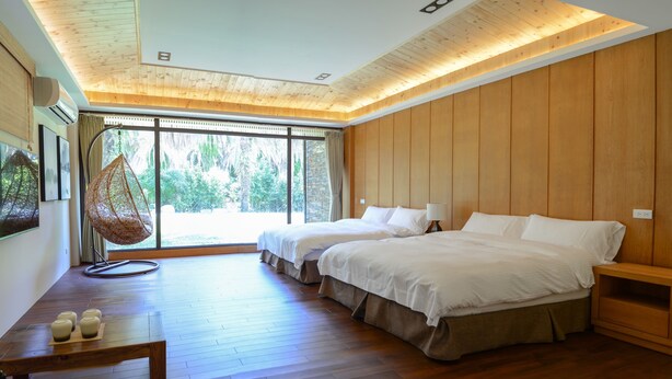 Quarto com duas camas, piso de madeira, uma cadeira suspensa e uma janela grande com vista para áreas verdes.