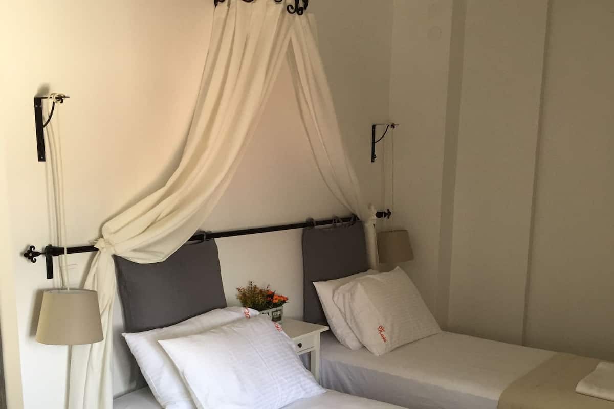 Une chambre d’hôtel avec deux lits simples, chacun doté de draps blancs et surmonté d’un baldaquin. Une petite table avec une lampe et un vase de fleurs se trouve entre les deux lits.