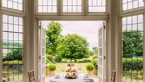 A table set for a tea service with a three-tiered tray of pastries, two glasses of water, and a teapot, positioned in a room with large windows overlooking a garden. - Falcon Manor Hotel (Settle)