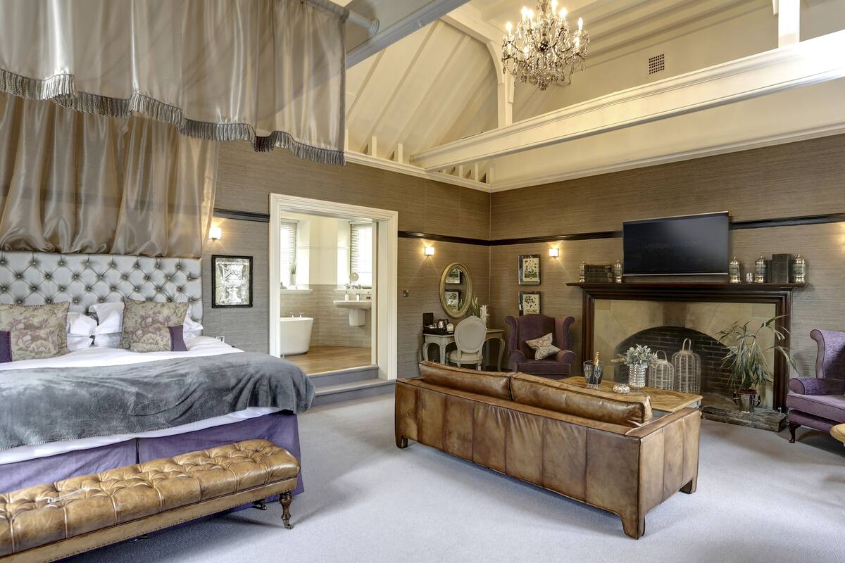 A spacious bedroom with a large bed, a leather bench, and a fireplace.