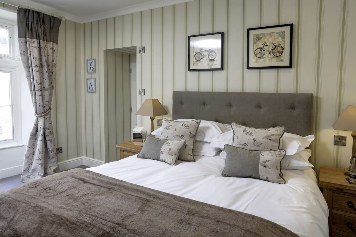 A neatly made bed with patterned pillows, a headboard, and a bedside table with a lamp. There are two framed pictures on the wall and a window with curtains.