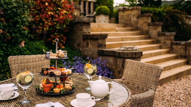 A wicker table set with a tiered dessert stand, a teapot, and two glasses of a beverage with a slice of cucumber. - Falcon Manor Hotel (Settle)