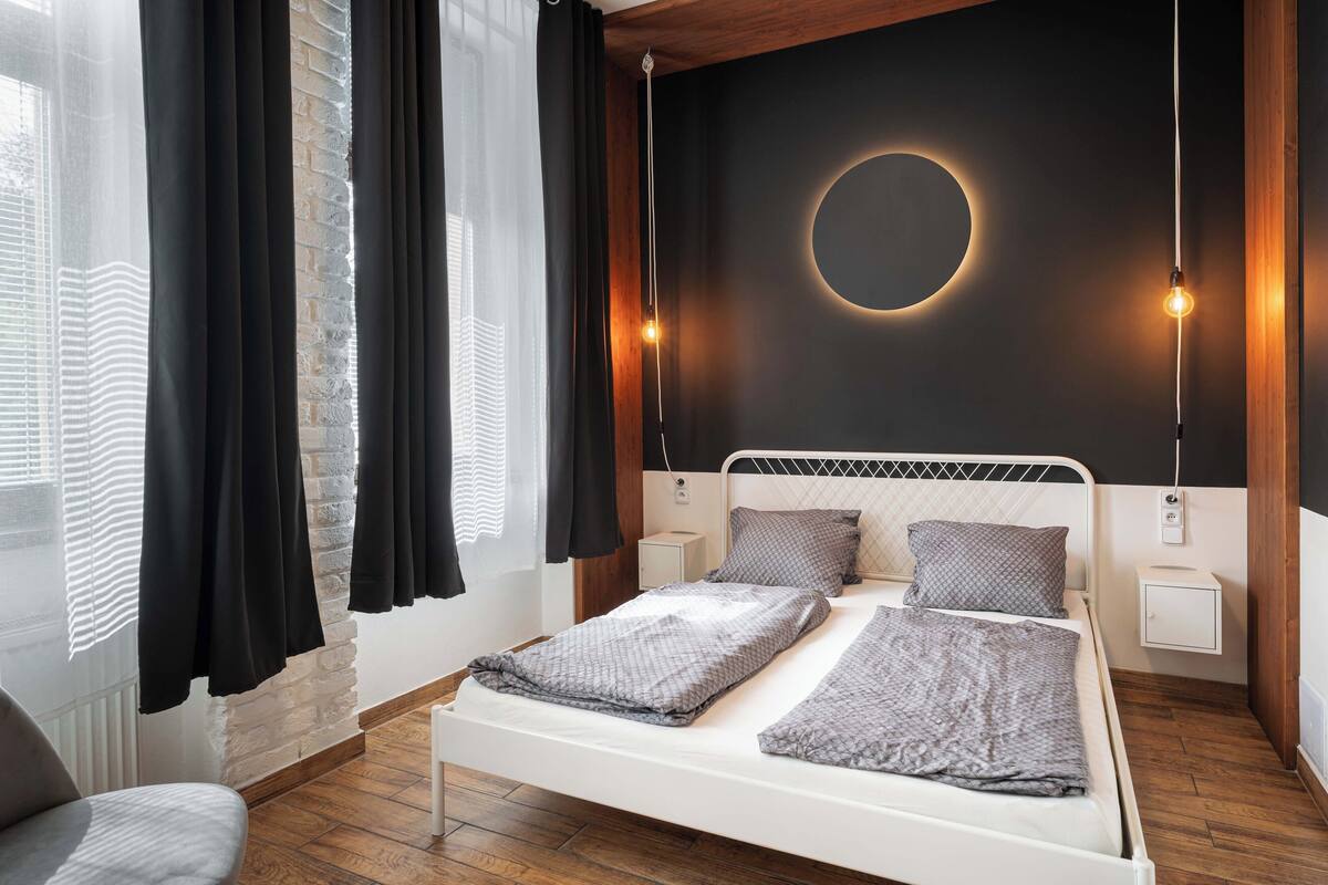 A modern bedroom with a bed, two bedside tables, a circular wall light, and a view of the city through large windows.
