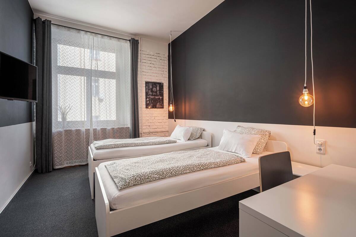A modern hotel room with two beds, a desk, and a large window with sheer curtains.