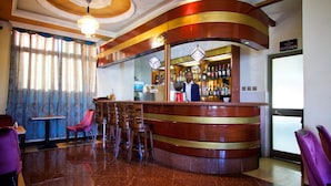 A bar area with a curved wooden counter, bar stools, and a bartender. There is a sign indicating the location of the kitchen. - Hera Addis Hotel (Addis Ababa)