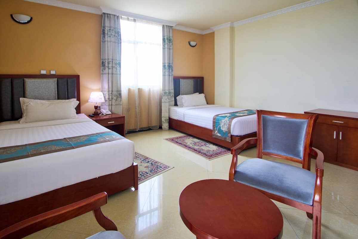 Quarto de hotel com duas camas, uma escrivaninha, uma cadeira e uma mesa de cabeceira com abajur e telefone.