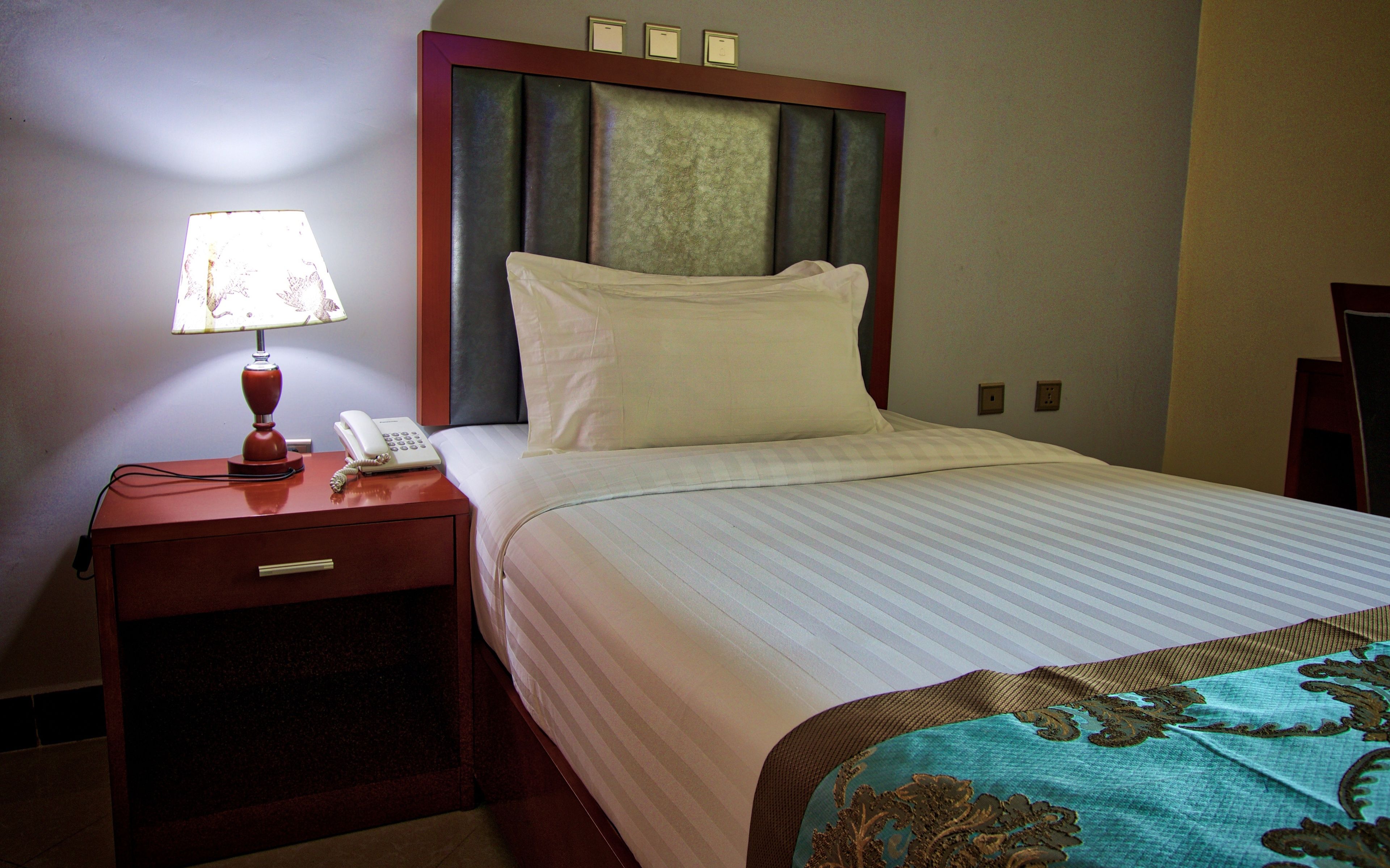 Habitación de hotel con cama, mesita de noche con lámpara y teléfono, y cabecera con panel decorativo.