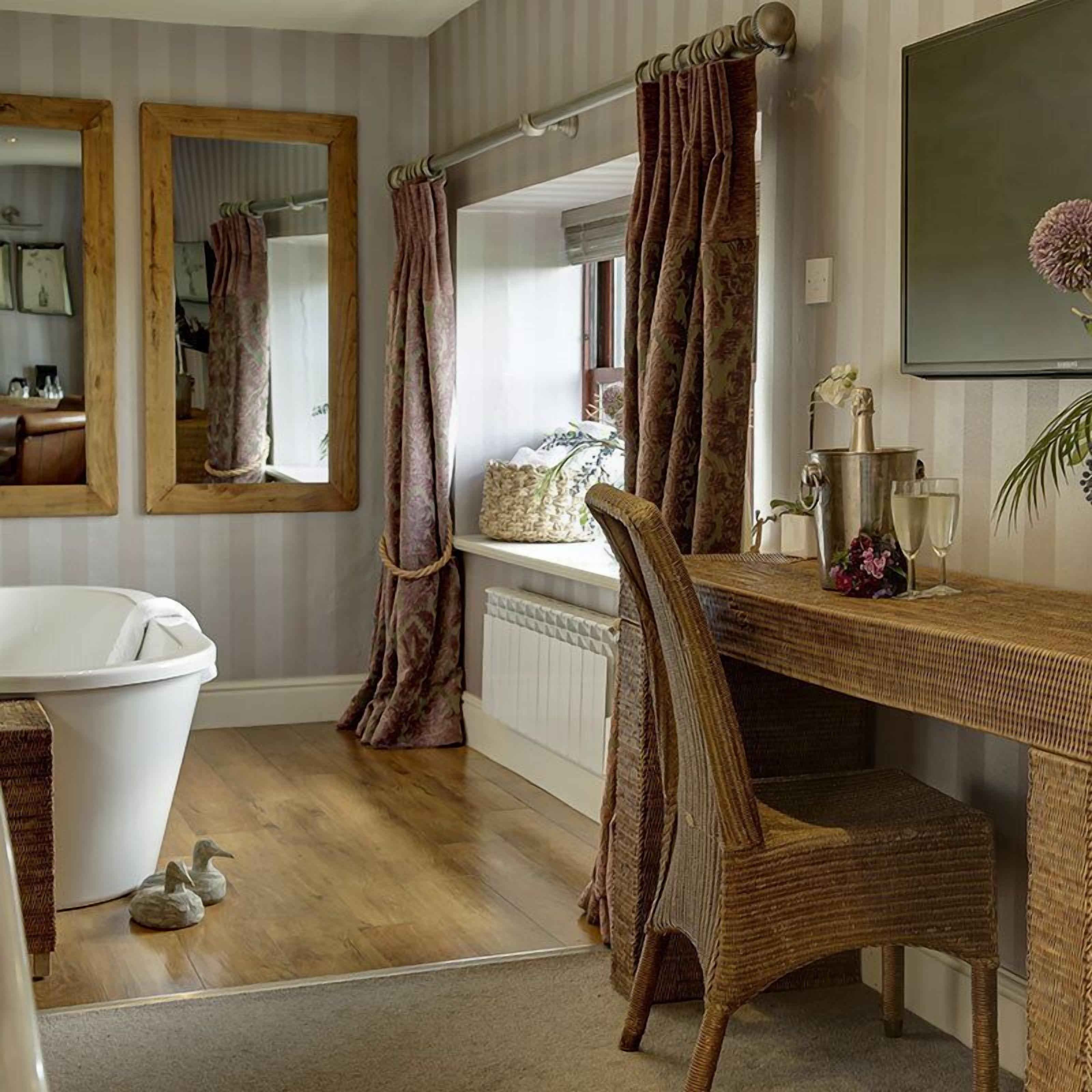 Un baño con bañera, una silla de mimbre, un tocador con espejo y una ventana con cortinas.