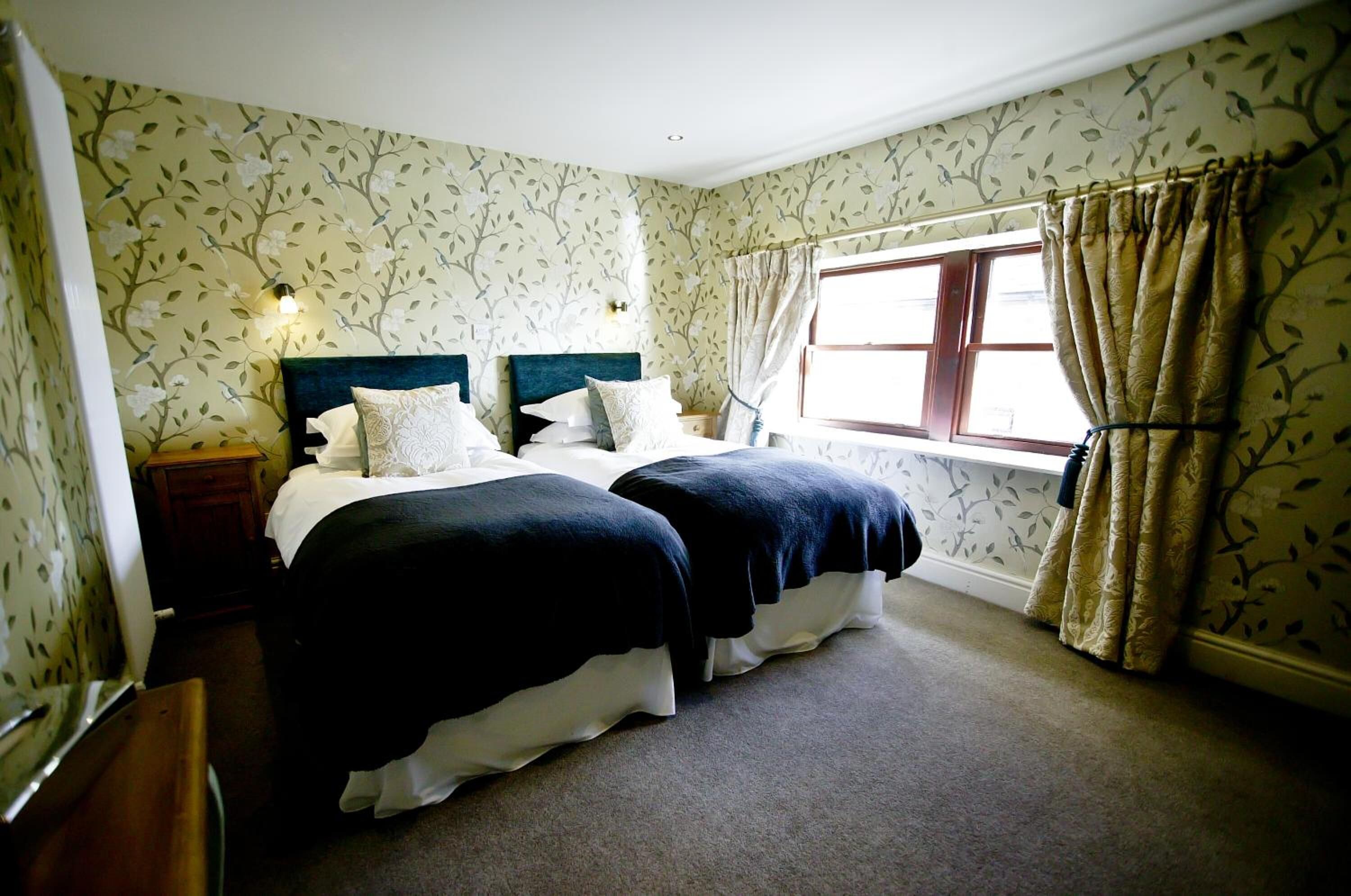 Un dormitorio con dos camas, papel tapiz floral, una ventana con cortinas y una mesita de noche.
