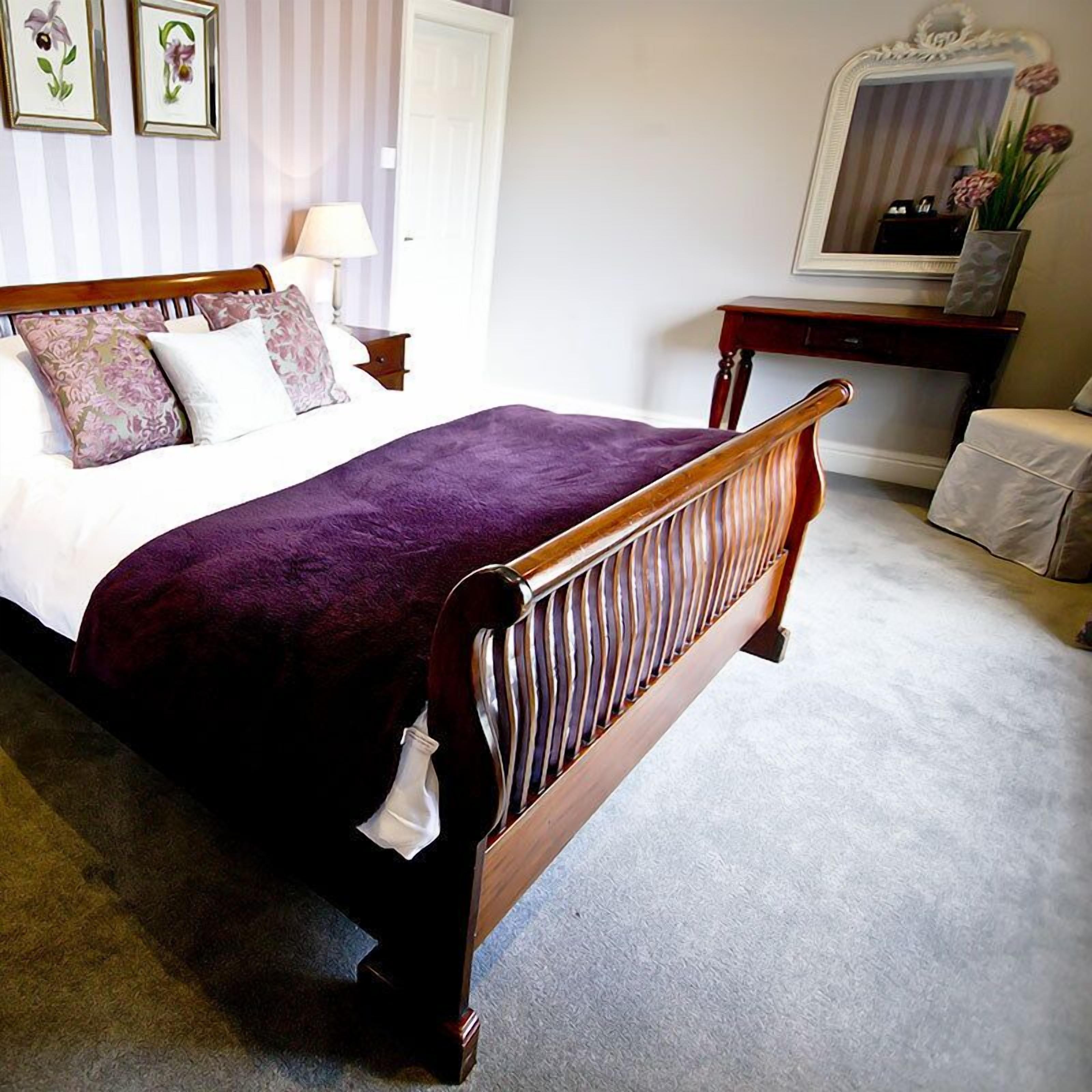 A bedroom with a wooden bed, a bedside table, a lamp, a mirror, and a small stool.