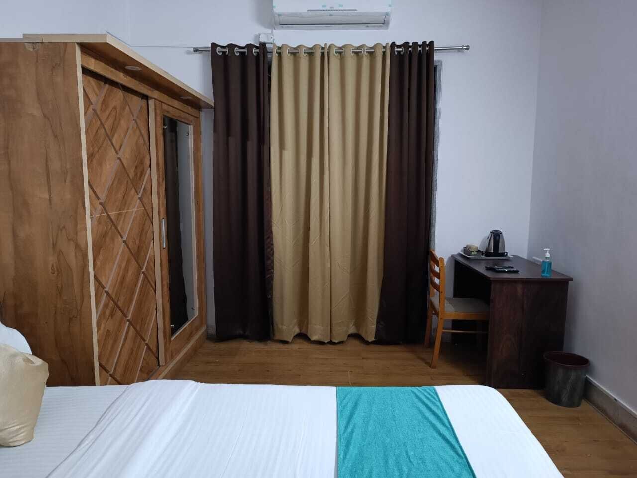 A bedroom with a bed, wooden wardrobe, curtains, a desk with a chair, and a trash bin.