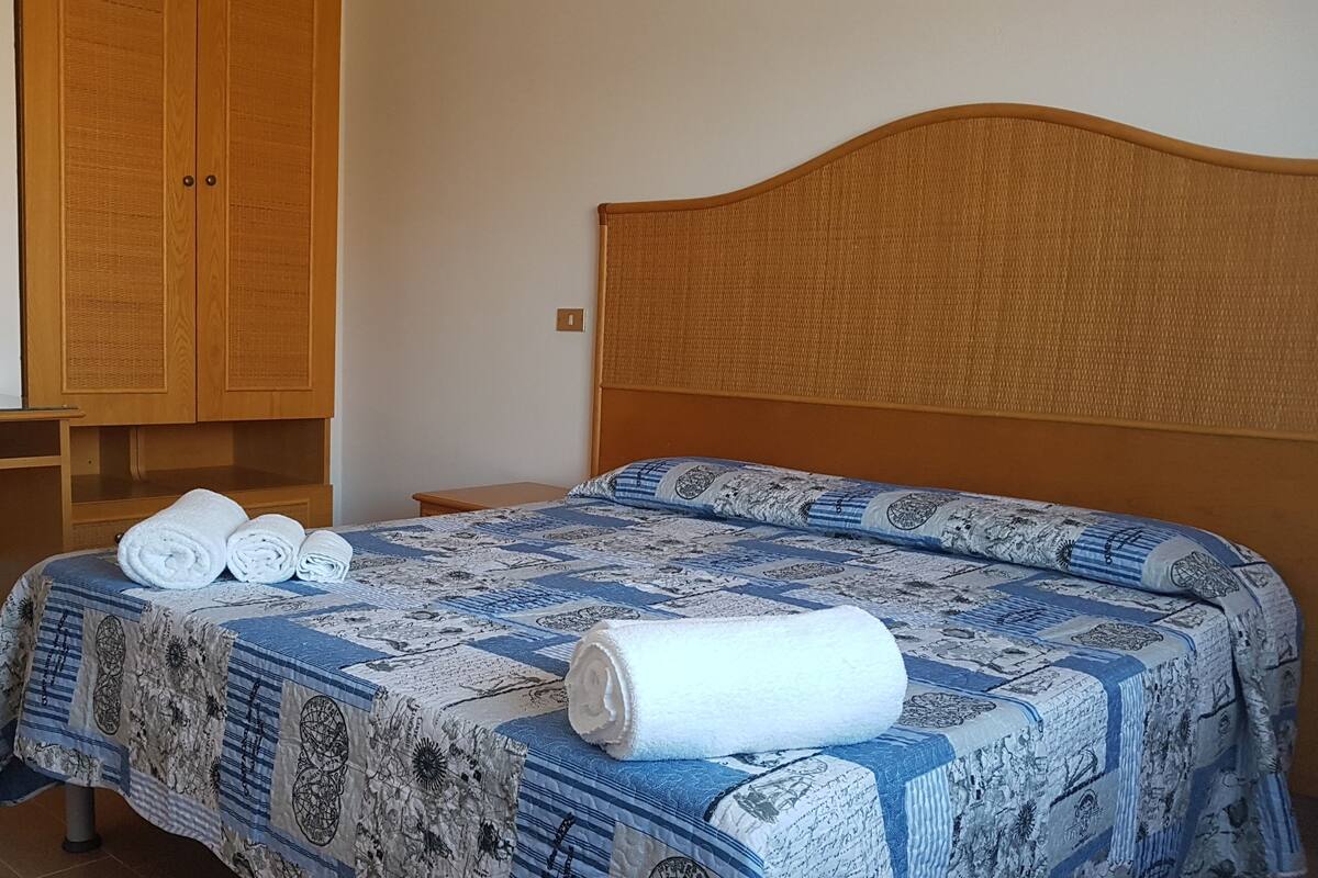A bedroom with a bed, a wooden wardrobe, a small bedside table, and a mirror.