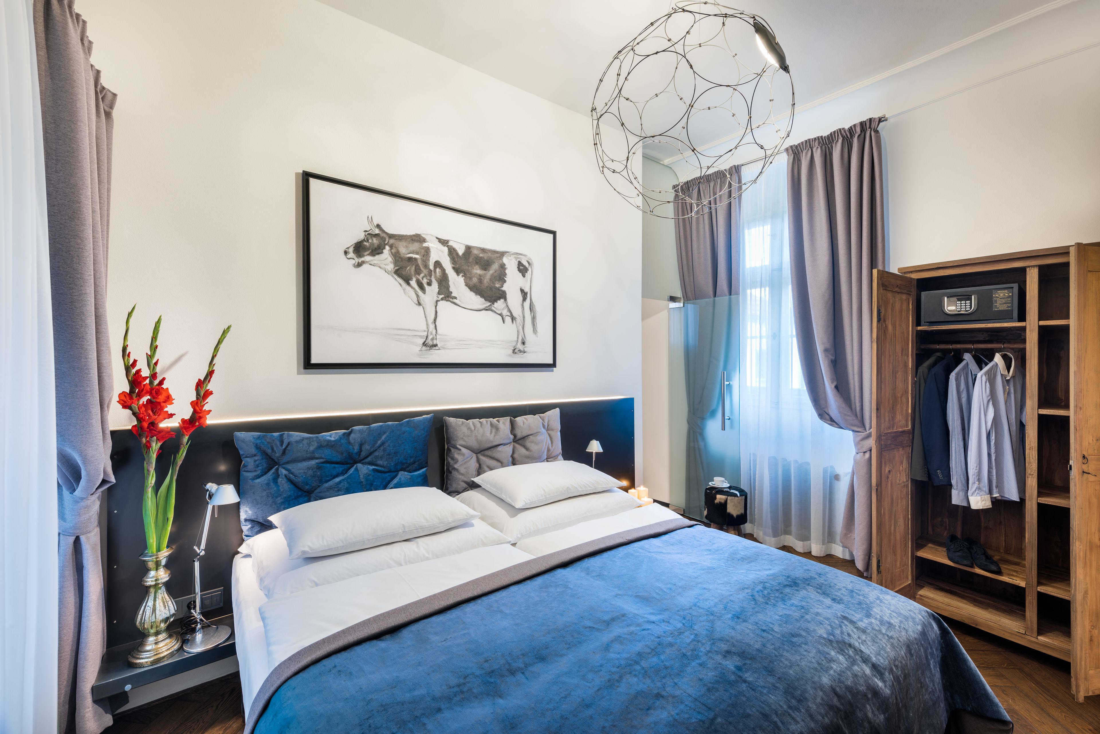 Une chambre moderne avec un grand lit, une table de chevet avec une lampe, une œuvre d’art représentant une vache accrochée au mur, et une armoire contenant des vêtements.