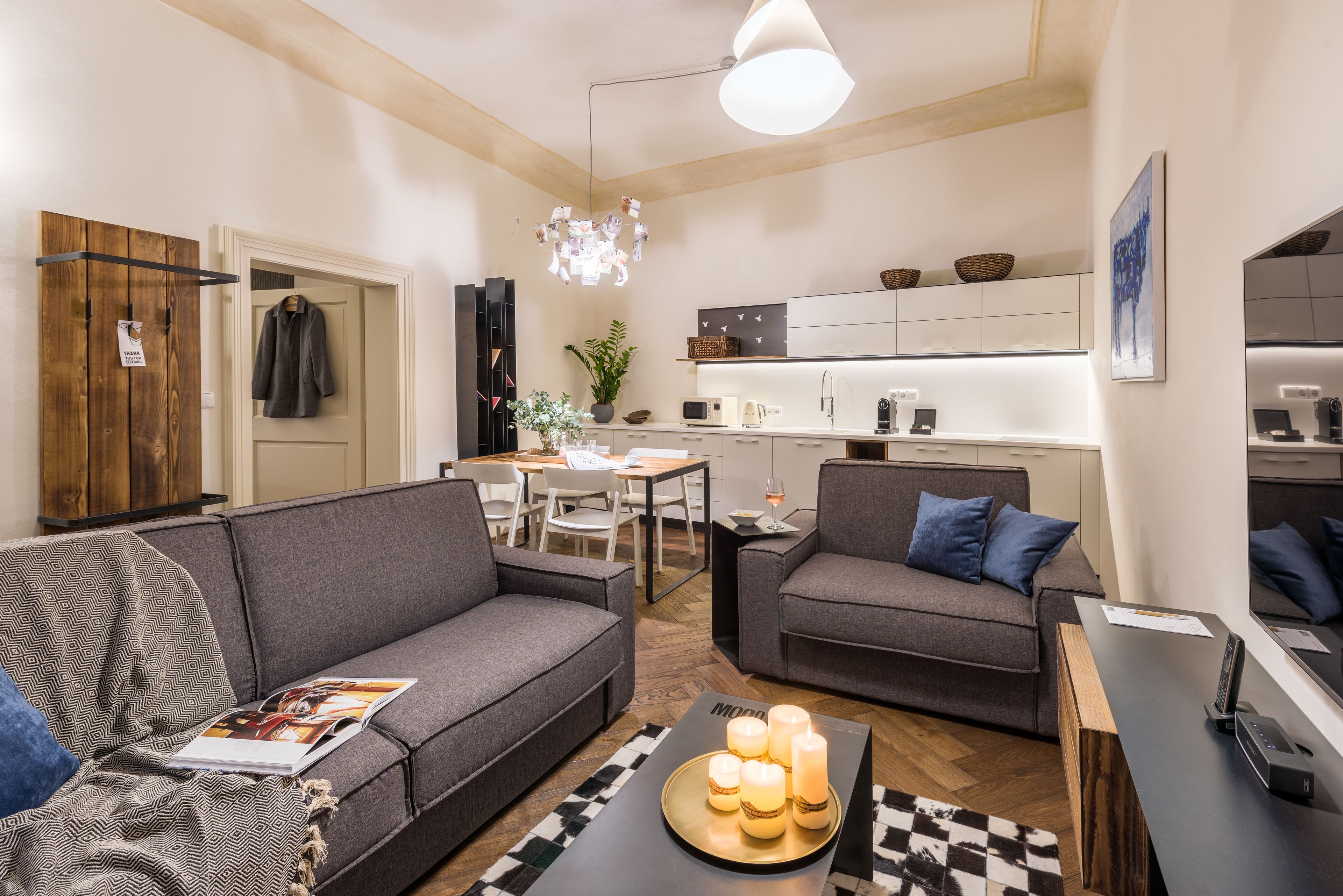 A modern living room with a sofa, armchair, and coffee table. There is a dining area with a table and chairs. The kitchen is visible with cabinets and appliances.