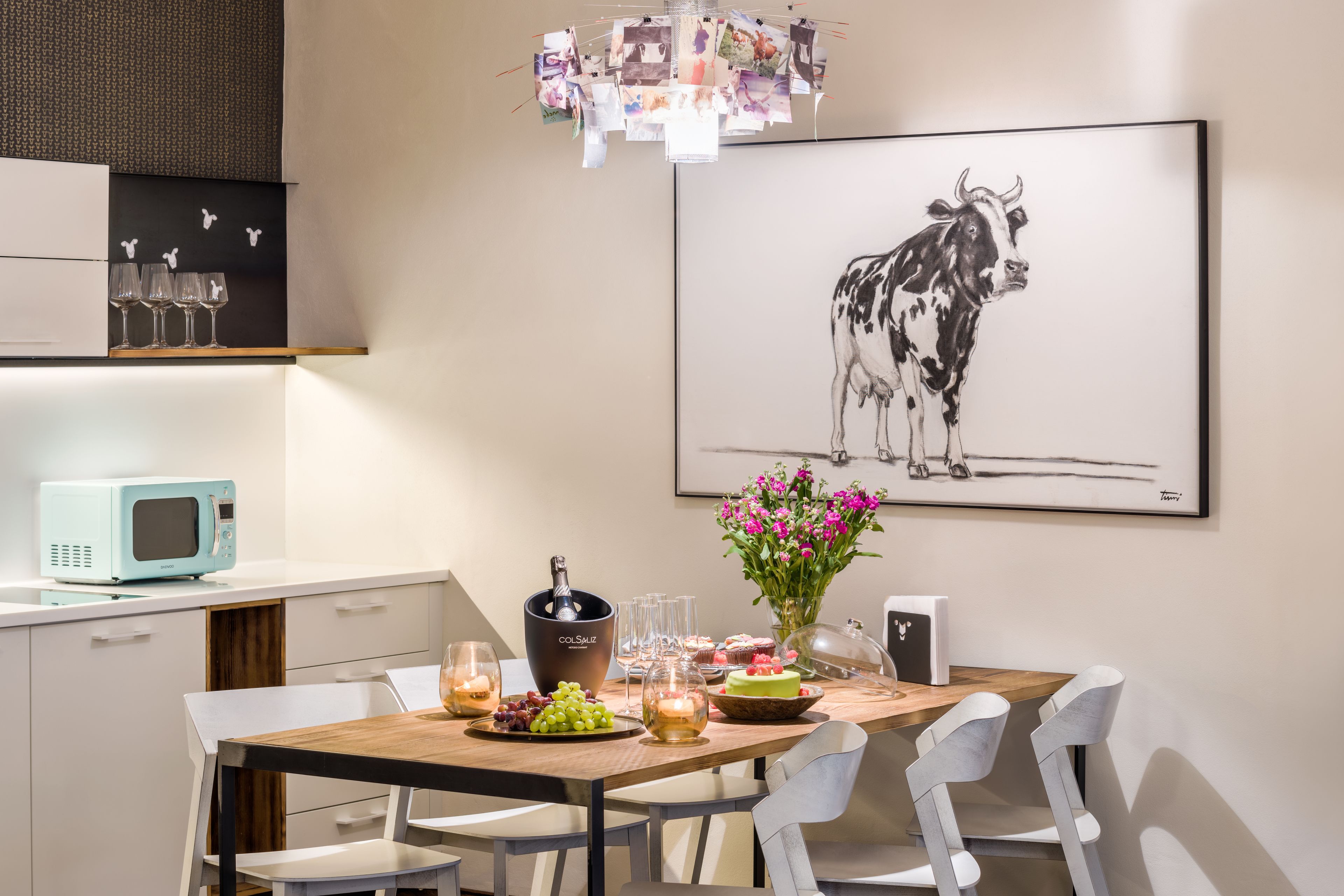 Un comedor con una mesa de madera, sillas blancas, un microondas azul y una obra de arte enmarcada de una vaca.
