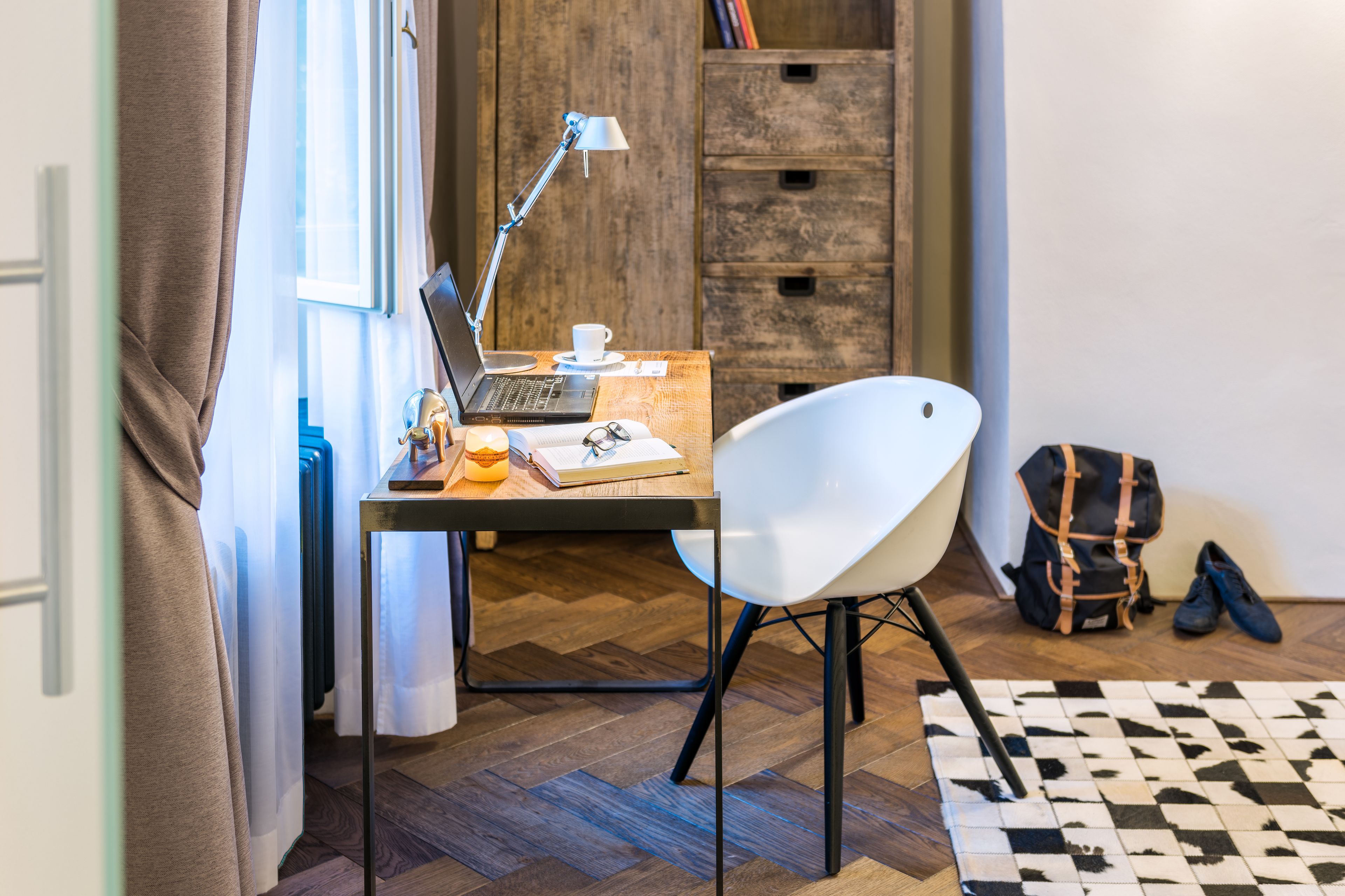 A room with a wooden herringbone floor, a desk with a laptop, a white chair, a lamp, a cup, a notebook, and a pair of shoes.