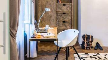 A room with a wooden herringbone floor, a desk with a laptop, a white chair, a lamp, a cup, a notebook, and a pair of shoes.