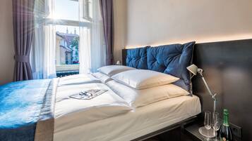 A neatly made bed with white and blue bedding, a bedside lamp, and a bottle of champagne on the nightstand.