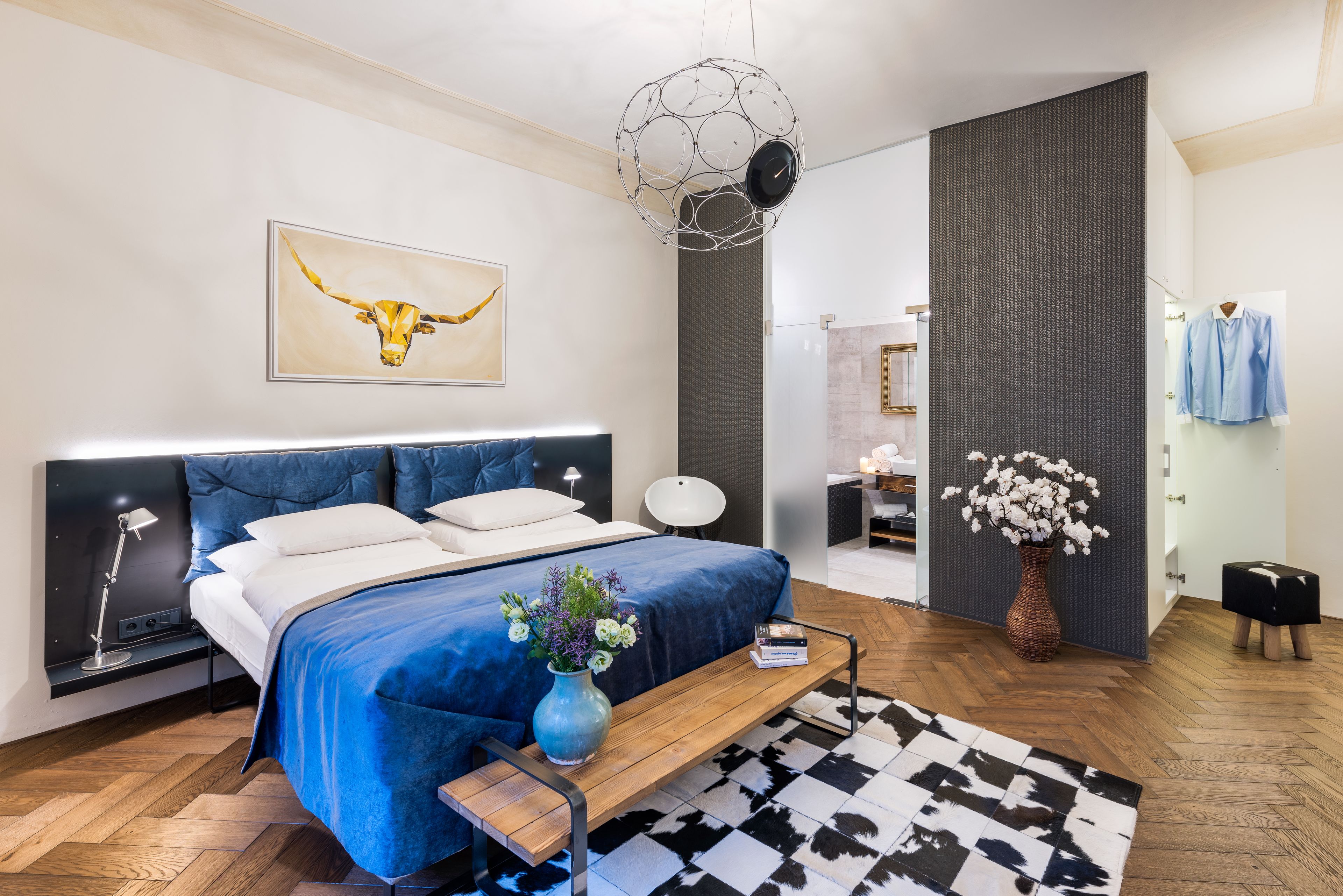 A modern bedroom with a large bed, a bedside table, a lamp, a wall-mounted artwork, a hanging light fixture, a mirror, and a small side table with a vase of flowers.