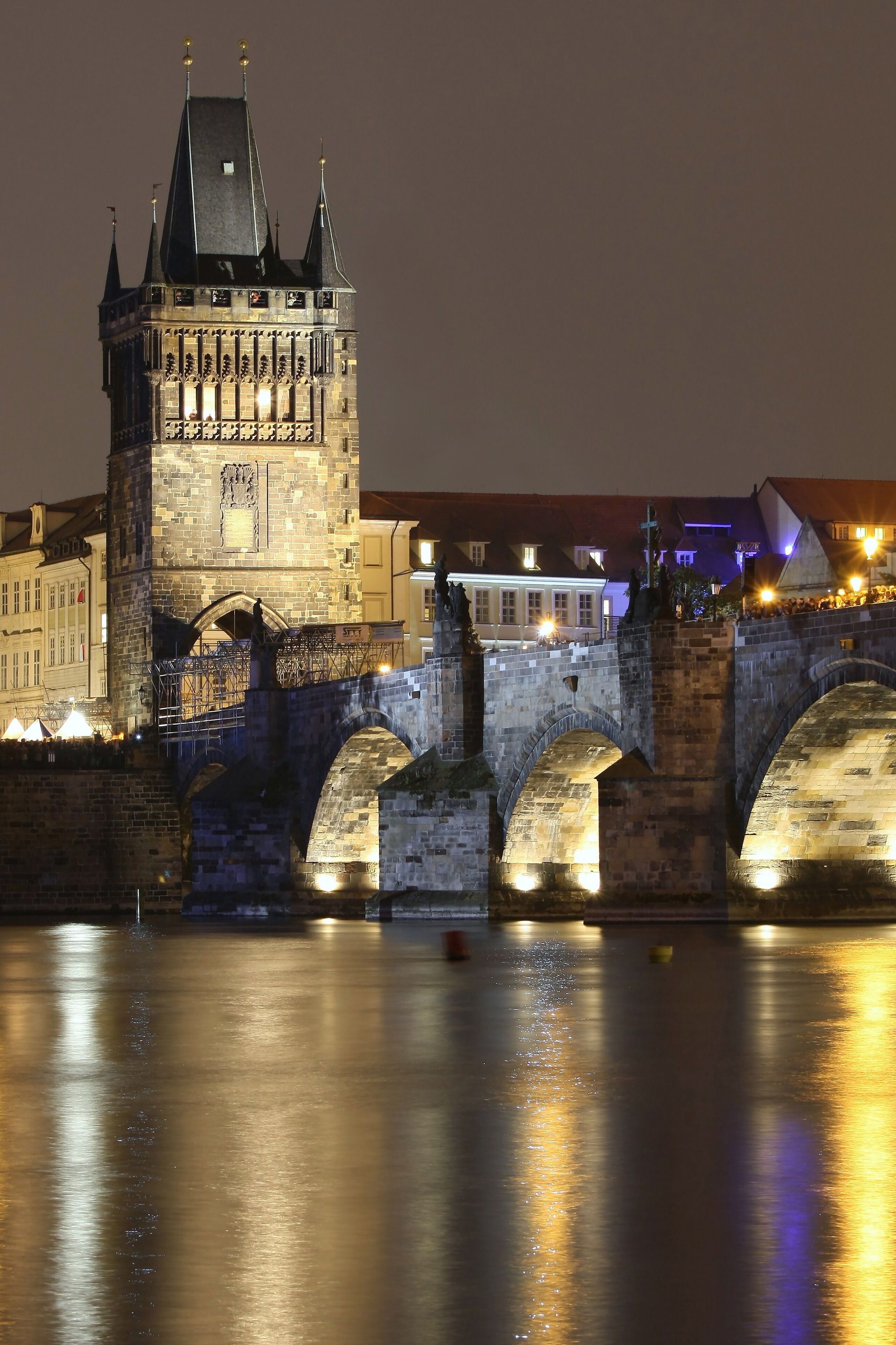 A historic stone bridge with arches, a tower with a pointed roof, and a calm river reflecting lights.