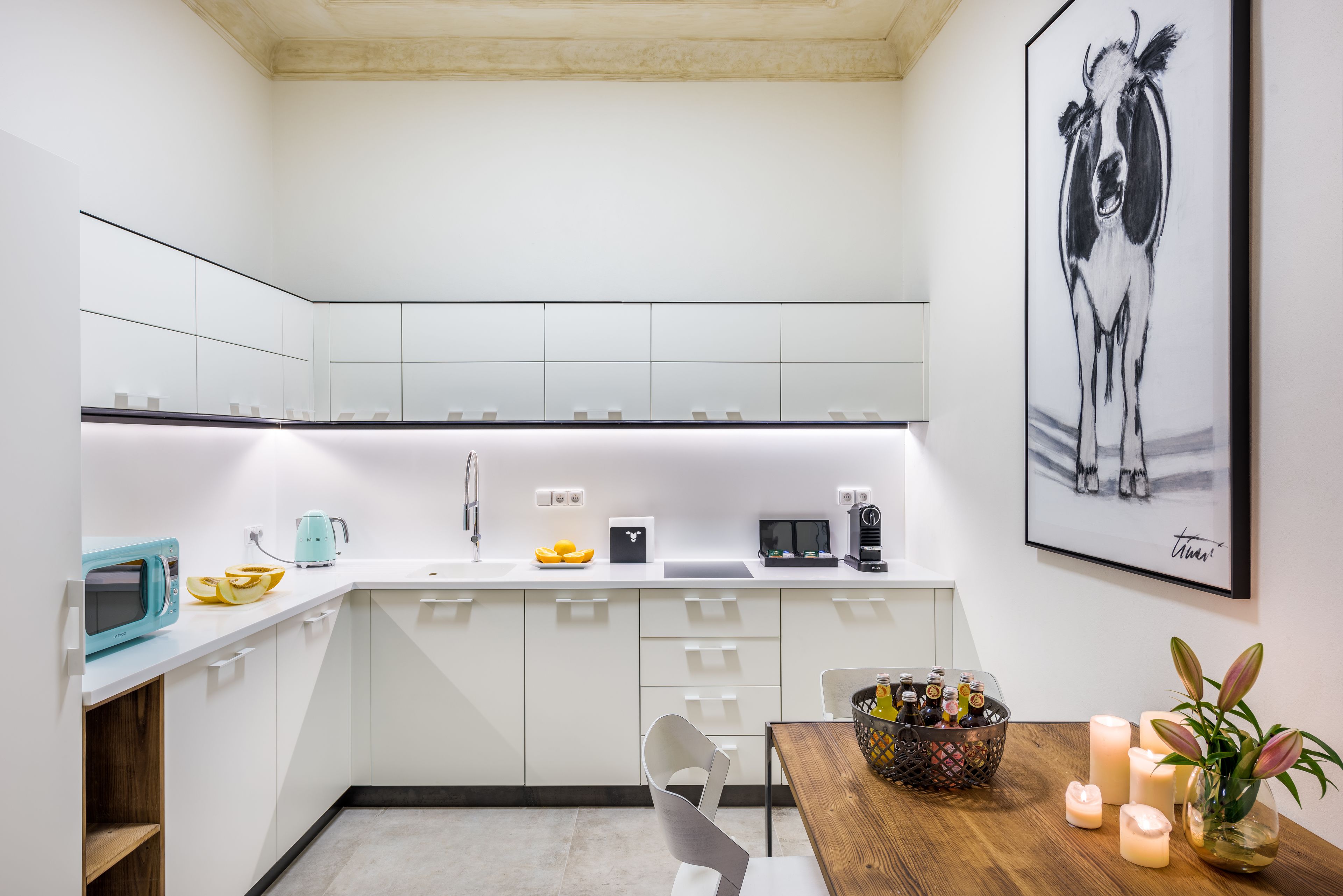 A modern kitchen with white cabinets, a microwave, a kettle, and a coffee maker. There is a framed artwork on the wall and a table with candles and a vase of flowers.