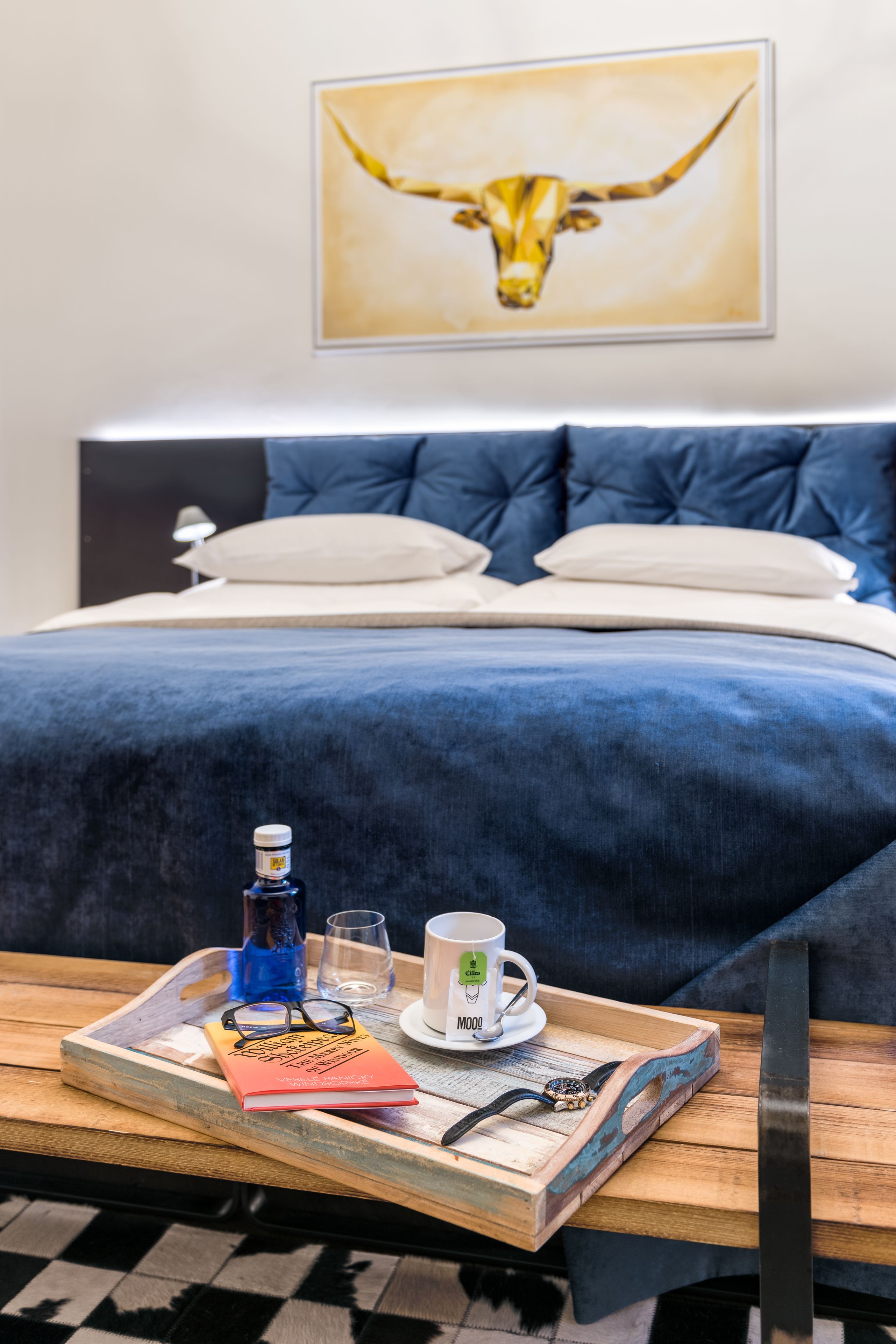 A neatly made bed with a blue bedspread, a tray with a book, glasses, a bottle, and a mug on a wooden table.