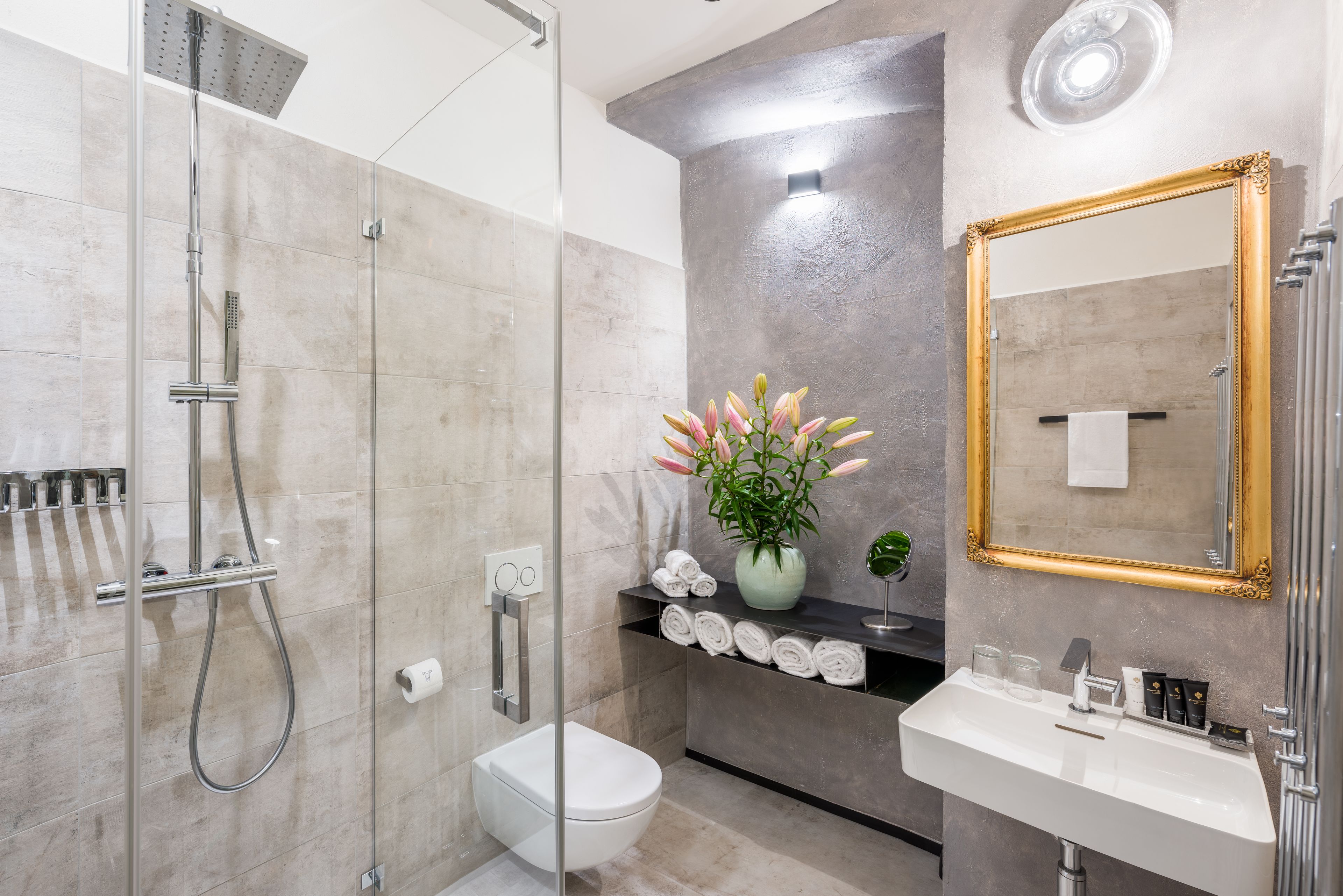 A modern bathroom with a glass-enclosed shower, a white toilet, a sink with toiletries, and a mirror with a decorative frame.