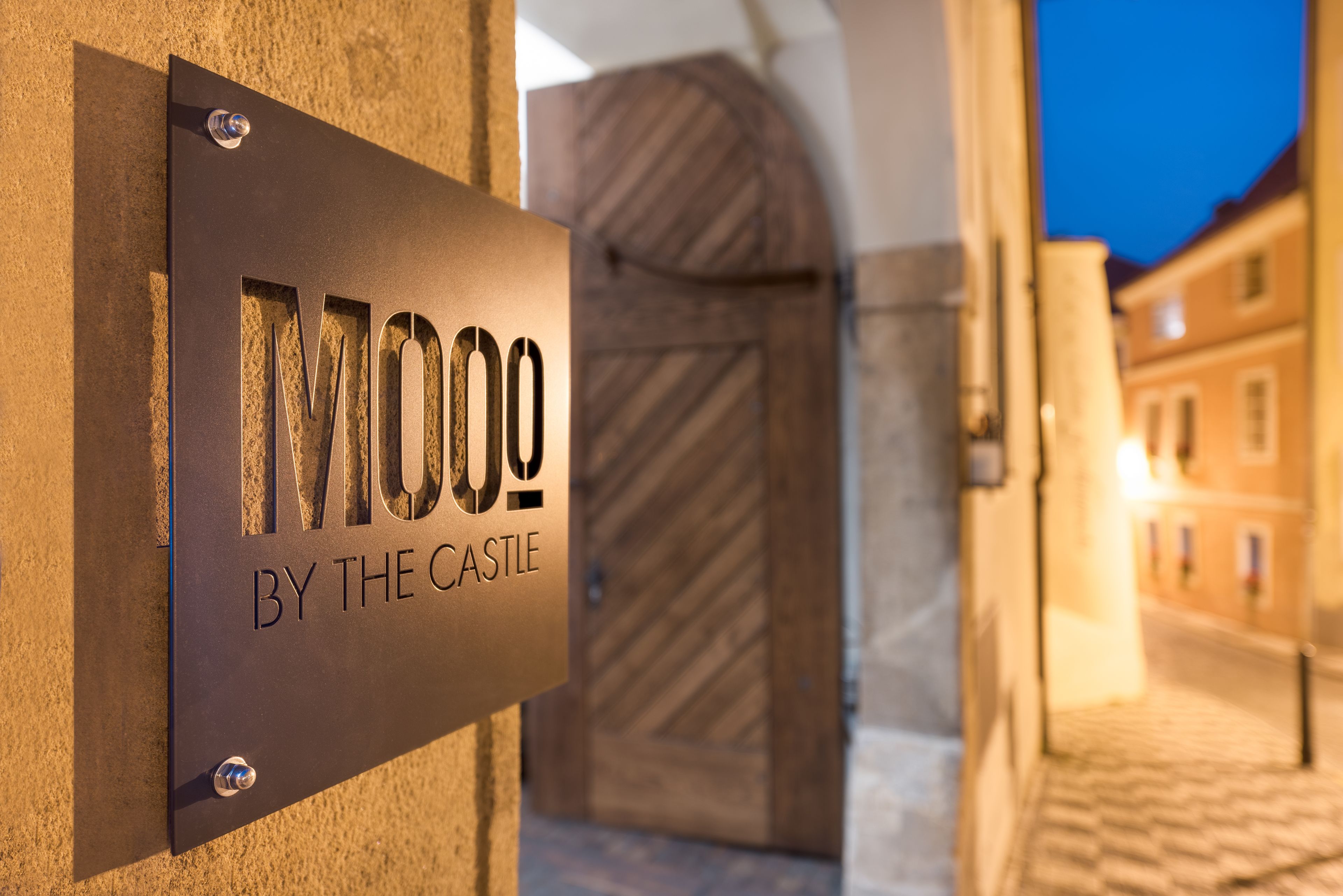 A sign with the text "MOOO BY THE CASTLE" mounted on a wall.