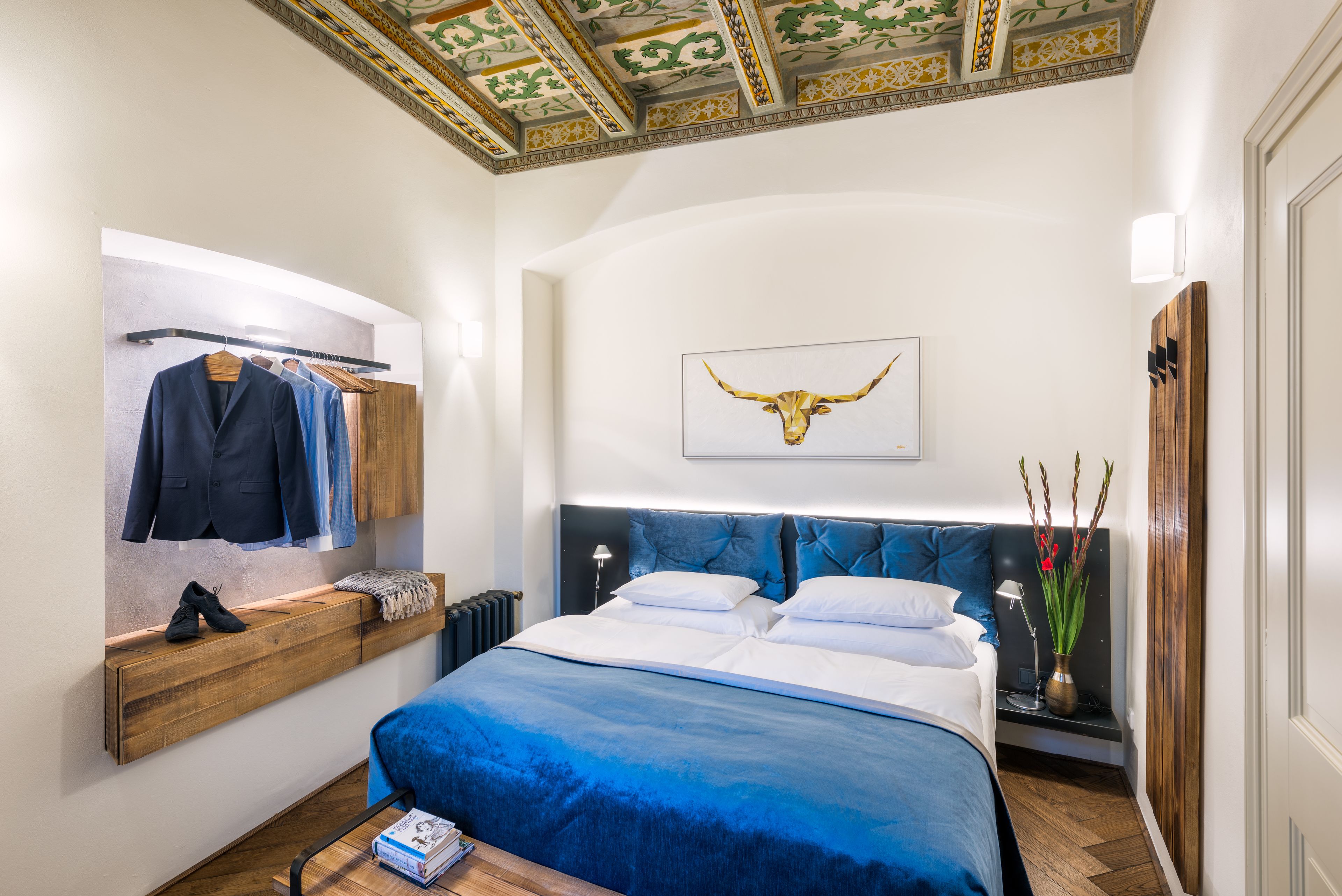 Un dormitorio con una cama grande, un estante de madera con ropa, un perchero, una obra de arte en la pared de una cabeza de toro y una mesita de noche con una lámpara y un florero.