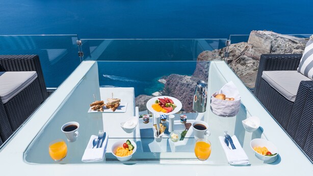 A table set with breakfast items, including fruits, bread, and beverages, overlooking a scenic coastal view. - Hyperion Oia Suites (Santorini)