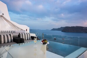A glass-top table with a wine glass and fruit bowl, overlooking a body of water with boats. - Hyperion Oia Suites (Santorini)