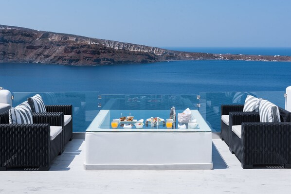 A terrace with a glass table set for breakfast, overlooking a cliff and the sea.