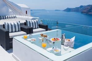 Una mesa con cubierta de vidrio, alimentos para el desayuno, un sofá de mimbre con cojines a rayas y vista al mar.