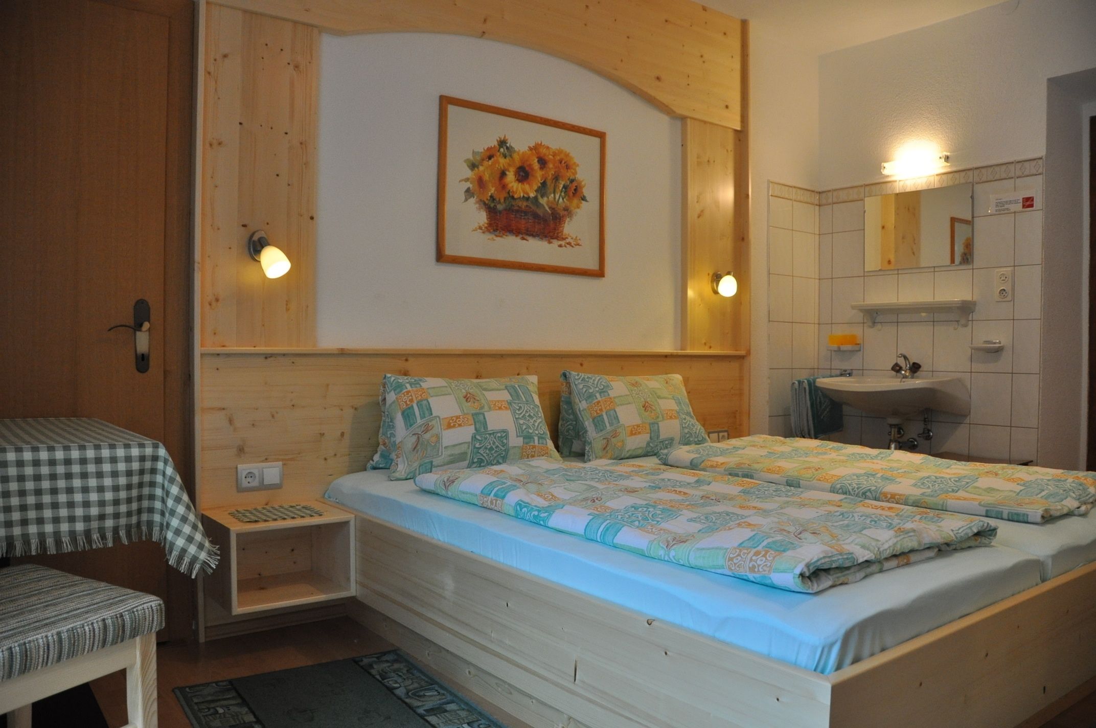 Ein Schlafzimmer mit Holz-Kopfteil, einem Bett mit gemusterten Textilien, einem Nachttisch, einer Bank, einer Wandleuchte und einem Spiegel über dem Waschbecken.