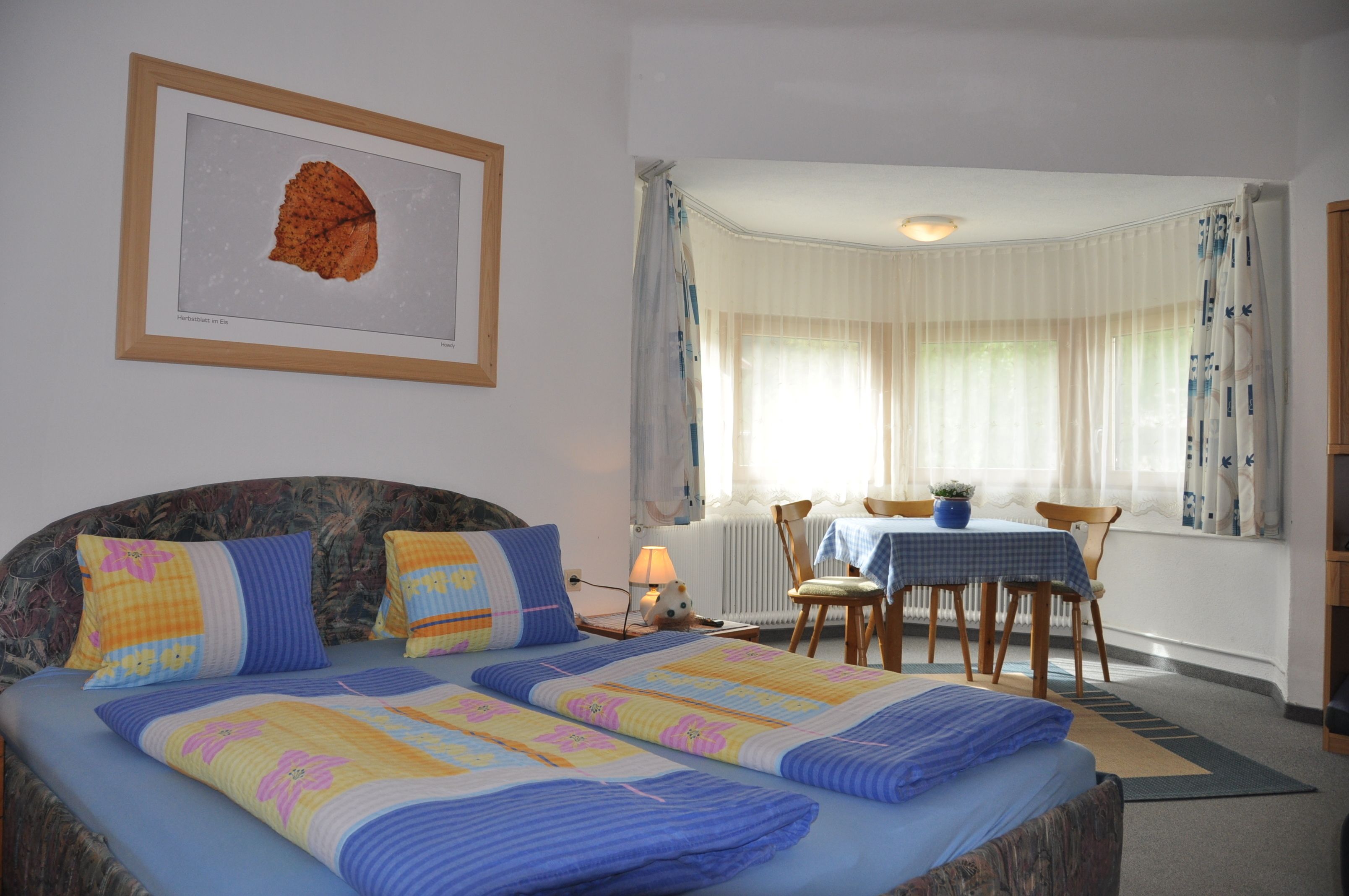 A bedroom with a bed, two pillows, a framed picture, a dining table with chairs, and a window with curtains.