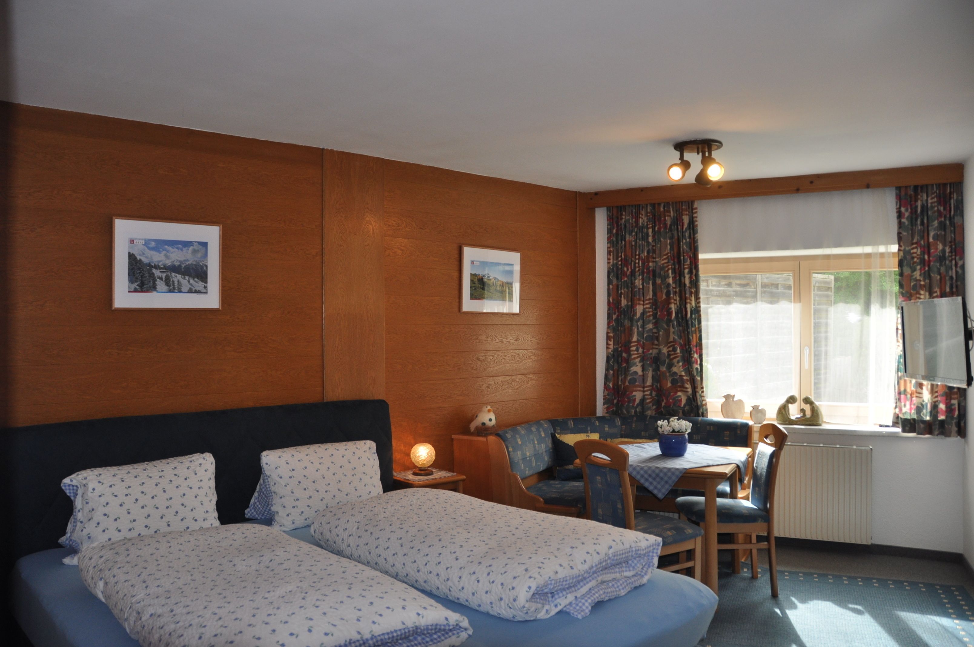 Ein Hotelzimmer mit einem Bett, einem kleinen Tisch mit Stühlen, einem Fenster mit Vorhängen und zwei gerahmten Bildern an der Wand.