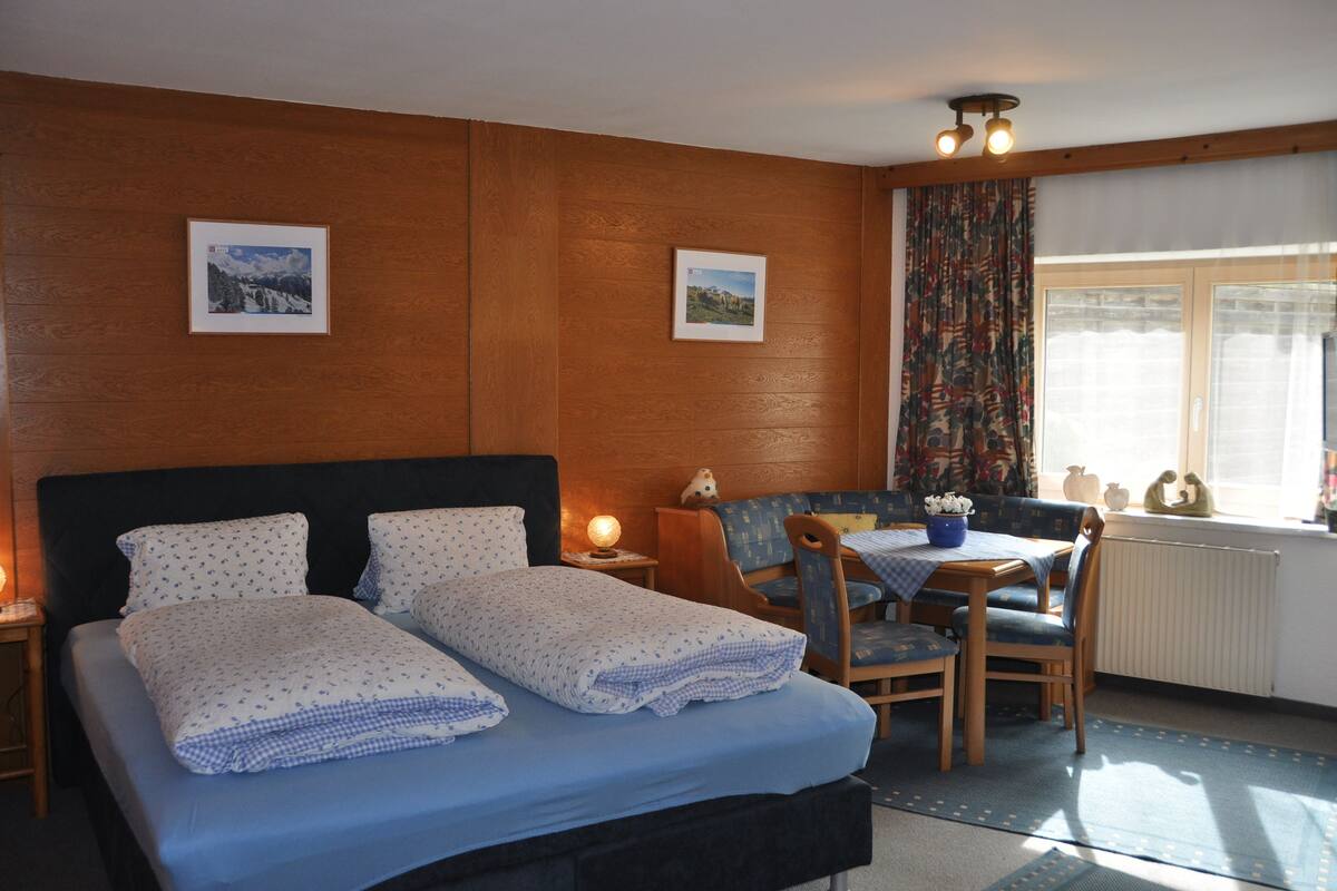 Een slaapkamer met een bed, nachtkastjes, een klein tafeltje met stoelen en een raam met gordijnen.