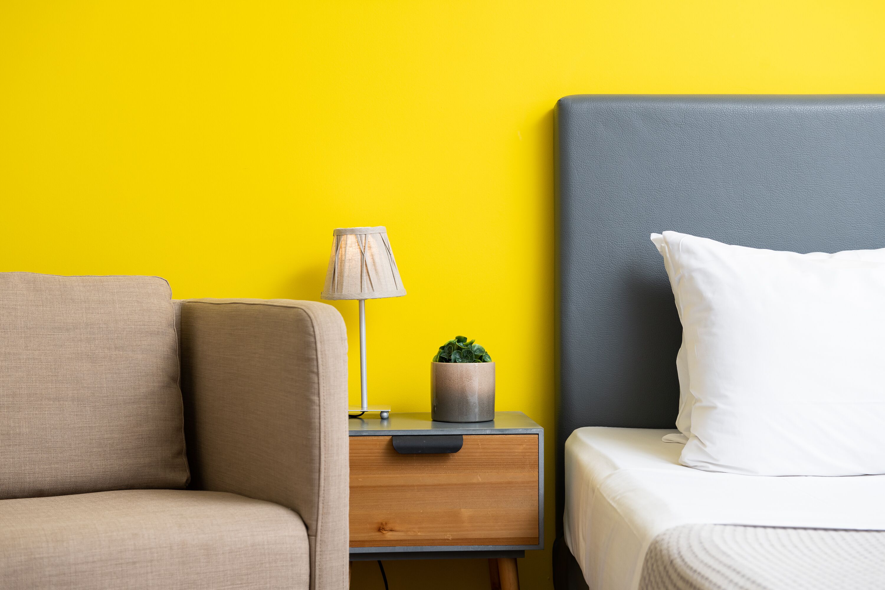 A room with a yellow wall, a grey headboard, a beige sofa, a wooden nightstand with a lamp and a plant, and a white pillow on the bed.