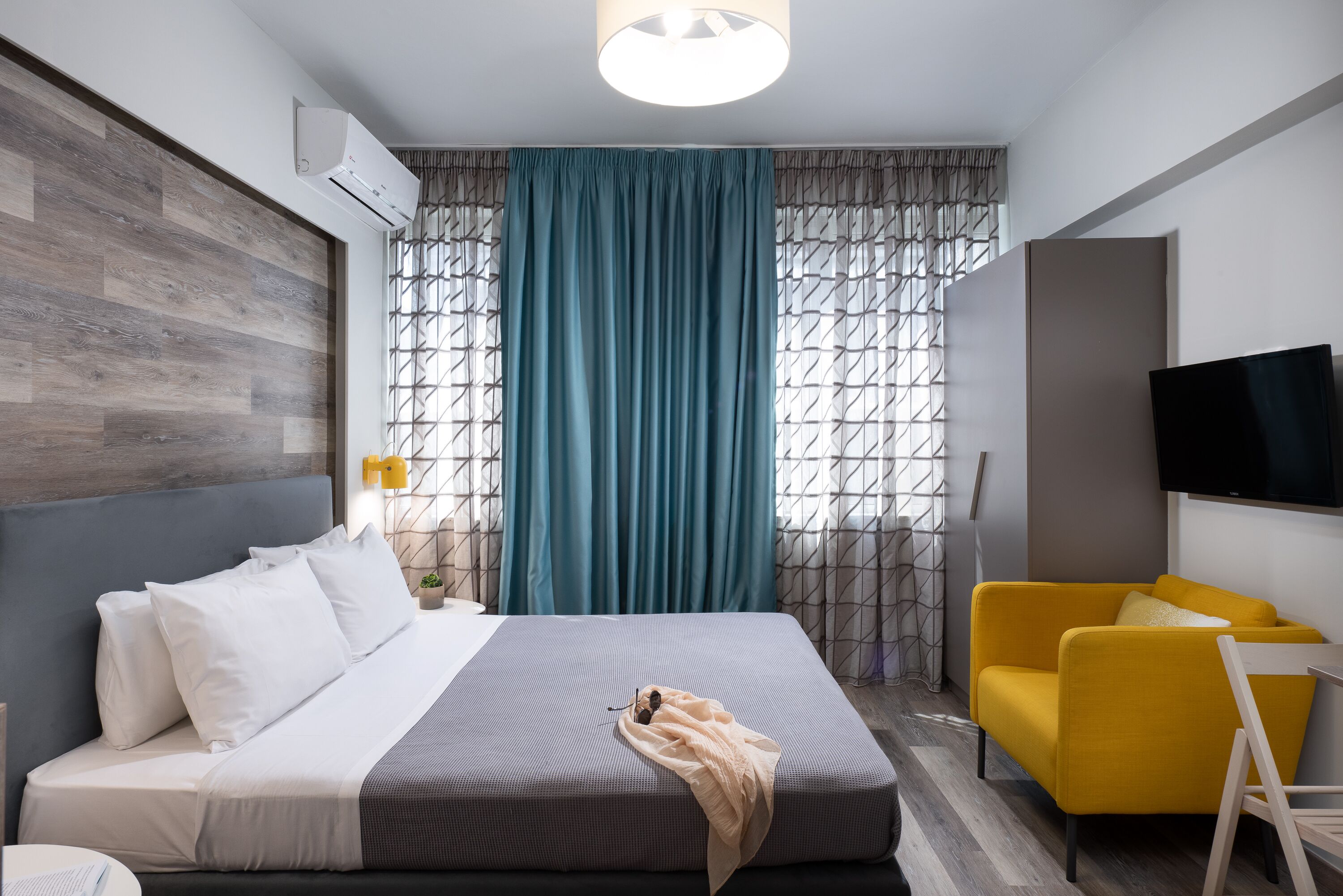 Une chambre d’hôtel moderne avec un lit, un fauteuil jaune, une télévision et une fenêtre avec des rideaux.