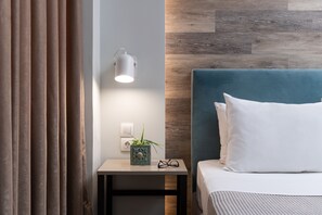 A modern bedroom with a blue headboard, white bedding, a small bedside table with a plant, and a wall-mounted light. - Enattica Syntagma Living (Athens)