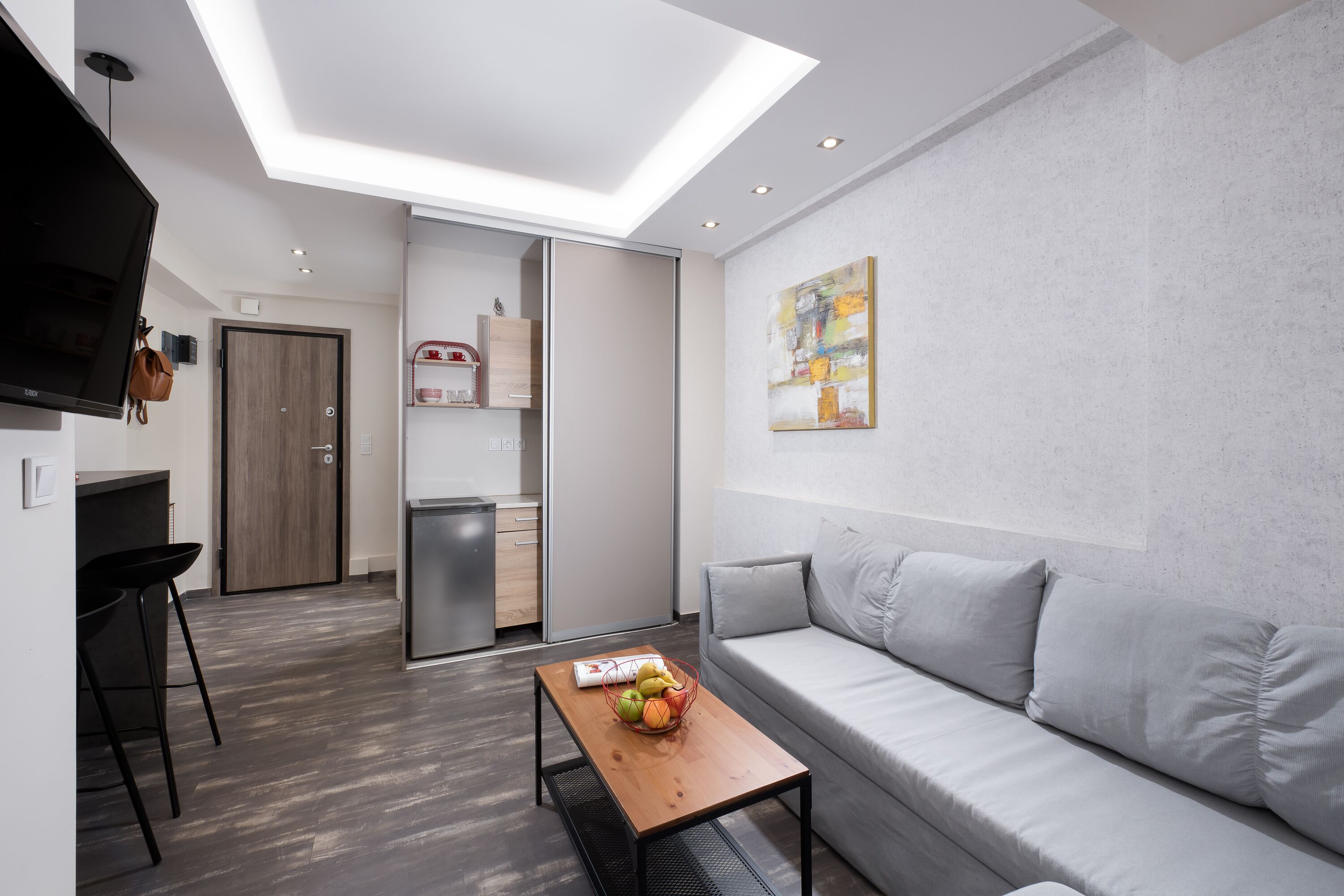 A modern living space with a sofa, a coffee table with fruits, a TV, and a kitchen area.