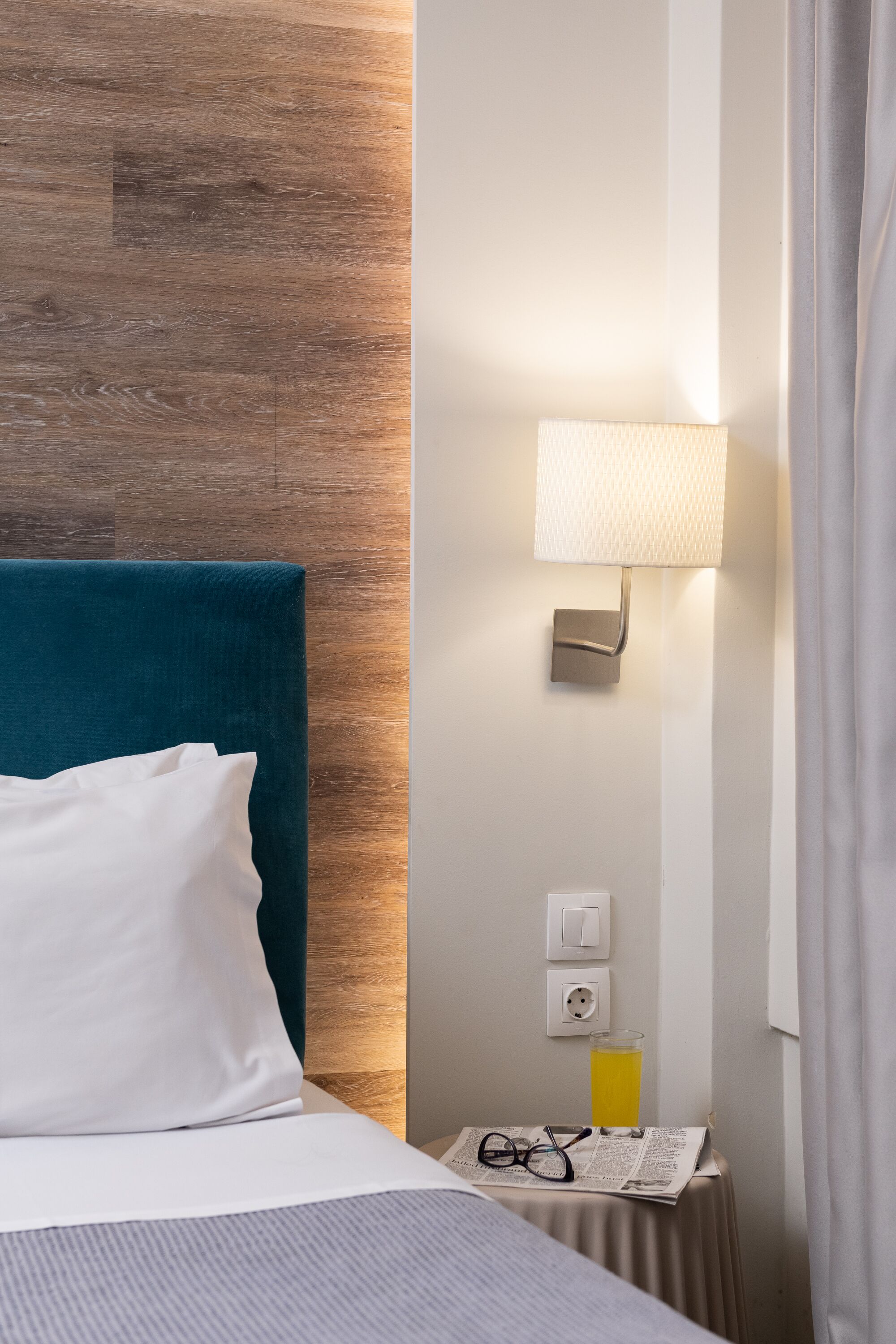 Quarto de hotel com cama, uma mesinha com abajur, um copo de suco e um jornal.