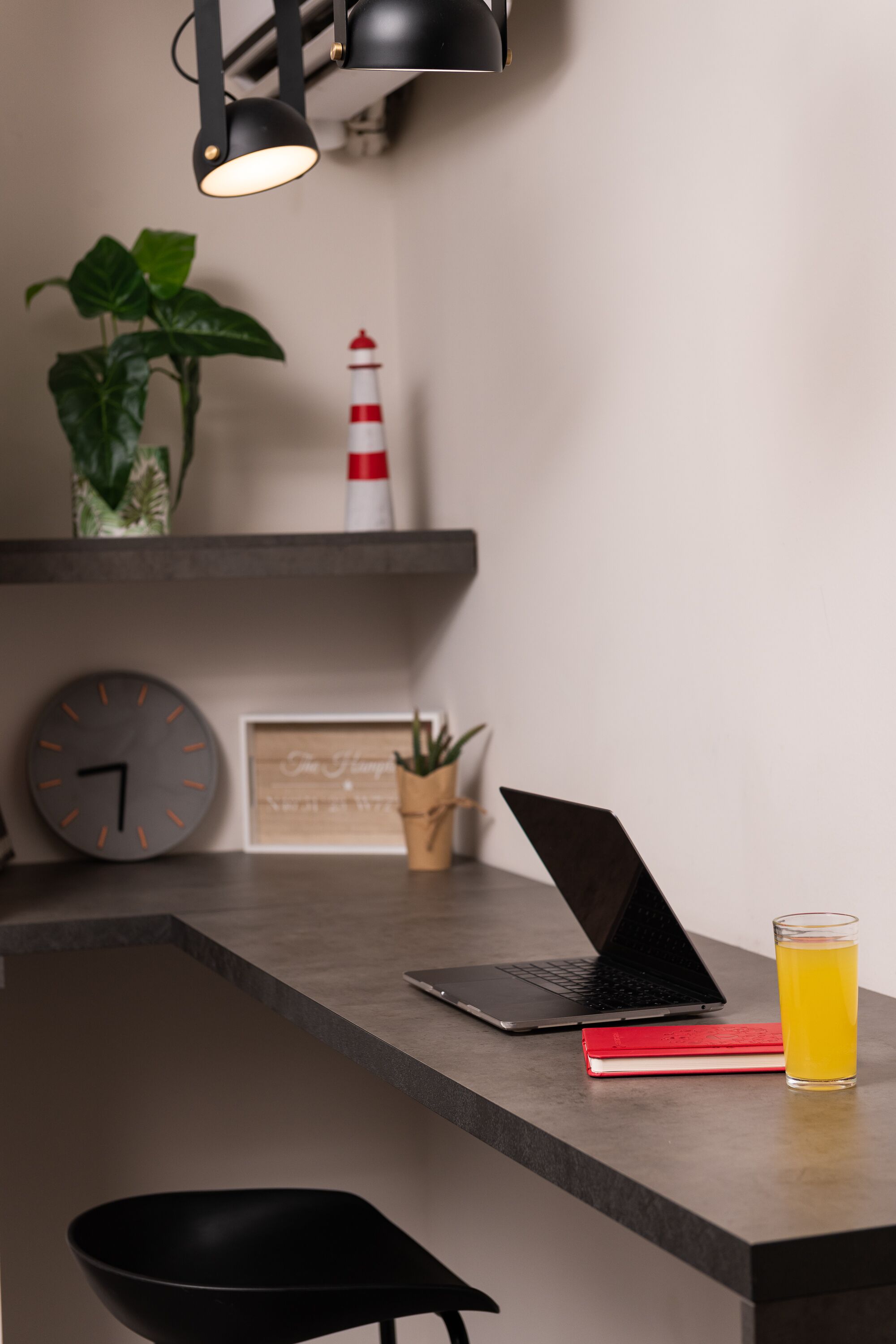 Een moderne keuken met een laptop, een glas sinaasappelsap, een rood boek, een zwarte stoel, een klok, een potplant en een model van een rode en witte vuurtoren.