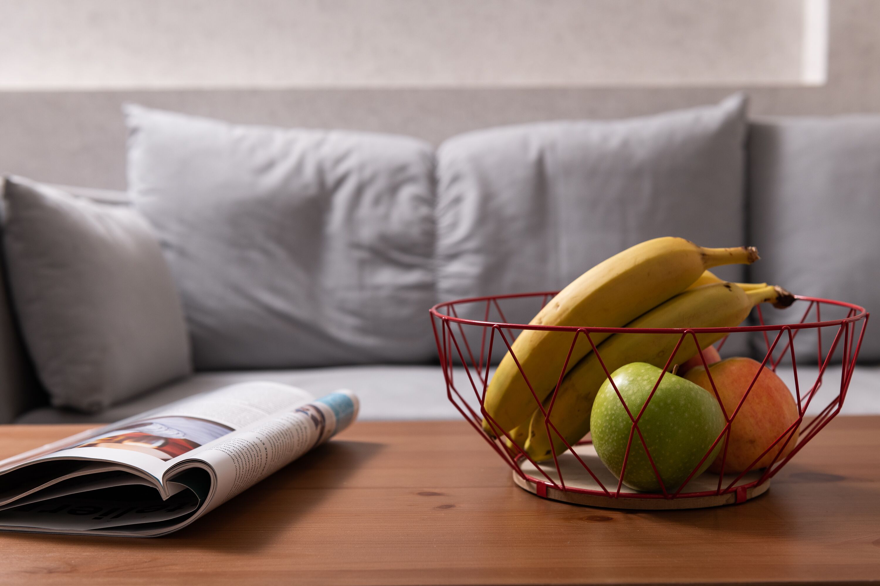 A wooden table with a fruit basket containing bananas, apples, and an orange. A newspaper is partially visible on the table.