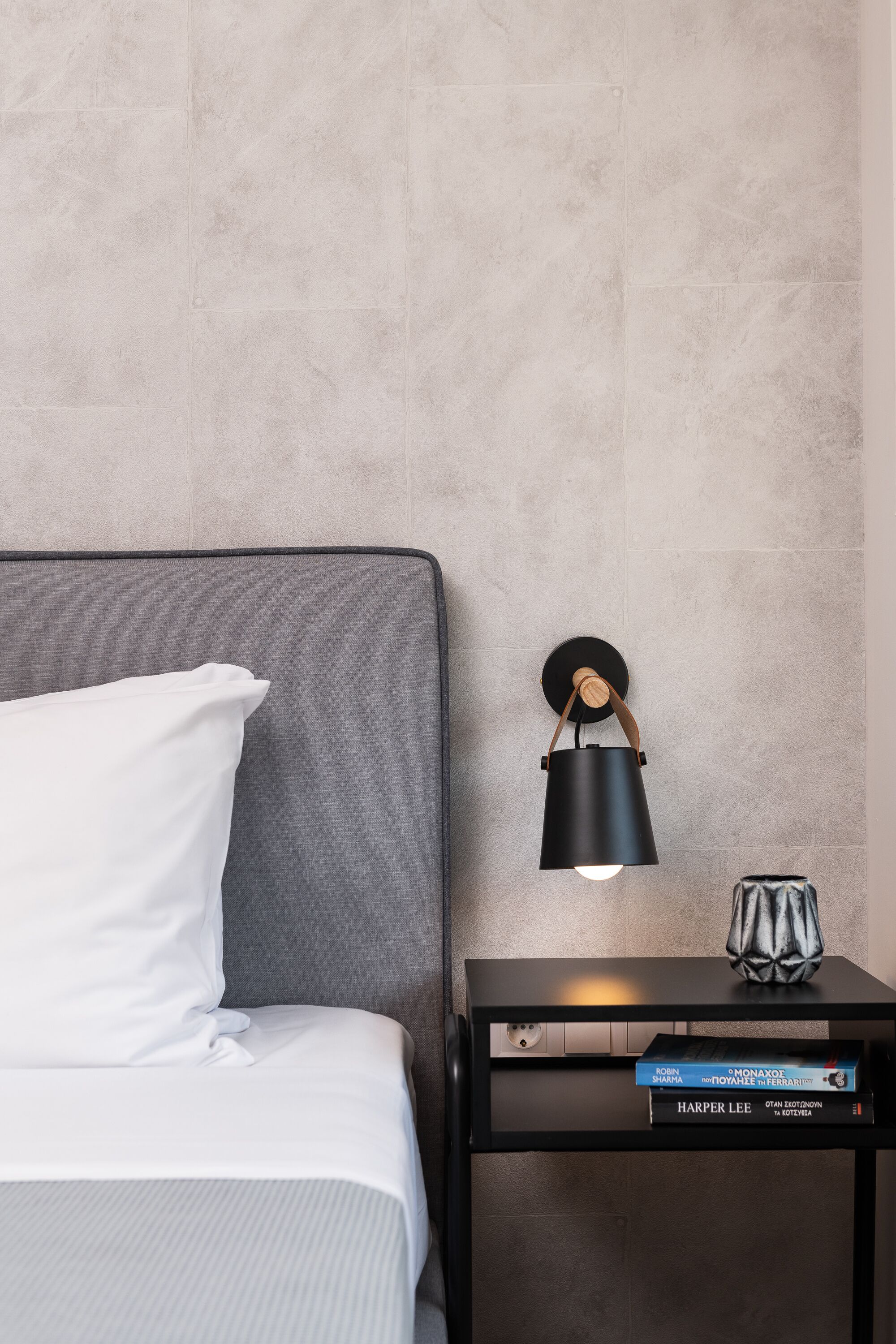 A neatly made bed with white bedding, a grey headboard, and a small bedside table with a black lamp and a few books.