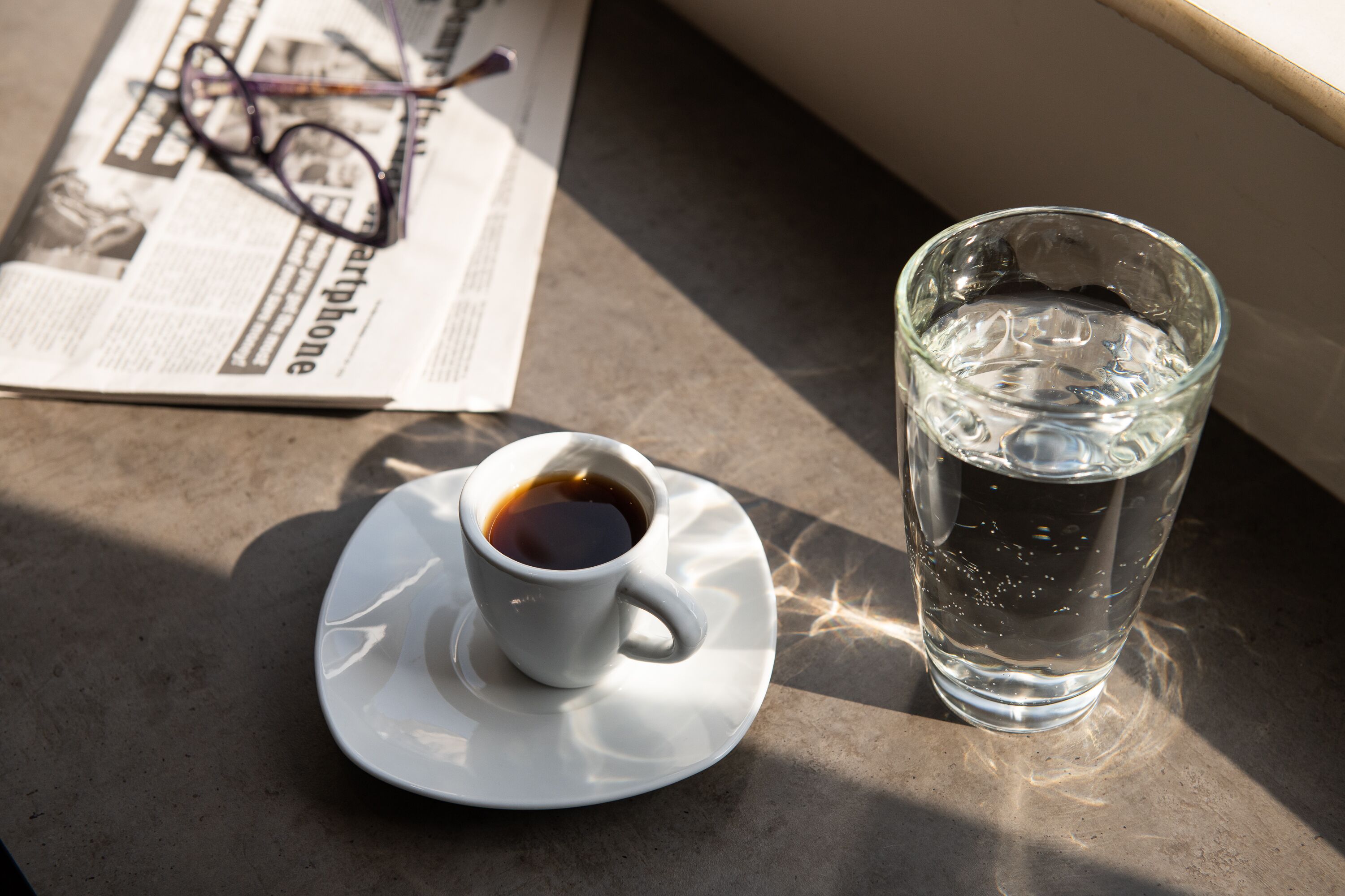 A cup of coffee and a glass of water on a table with a newspaper and glasses.