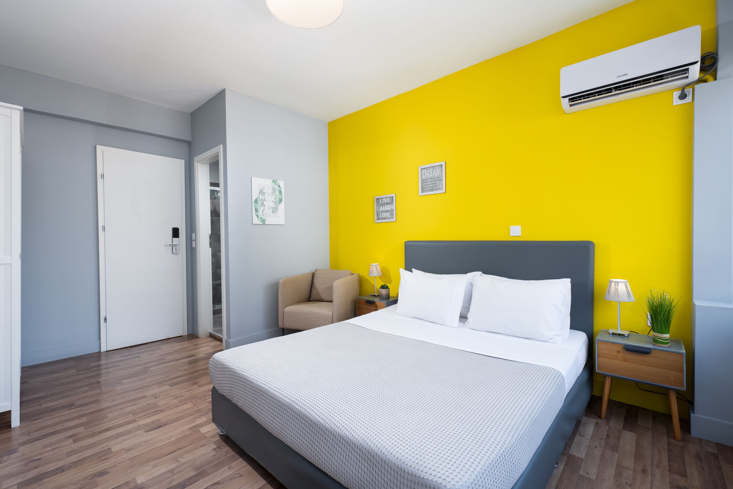 Una habitación de hotel moderna con una cama grande, una silla, una mesita de noche con lámpara y un equipo de aire acondicionado en la pared.