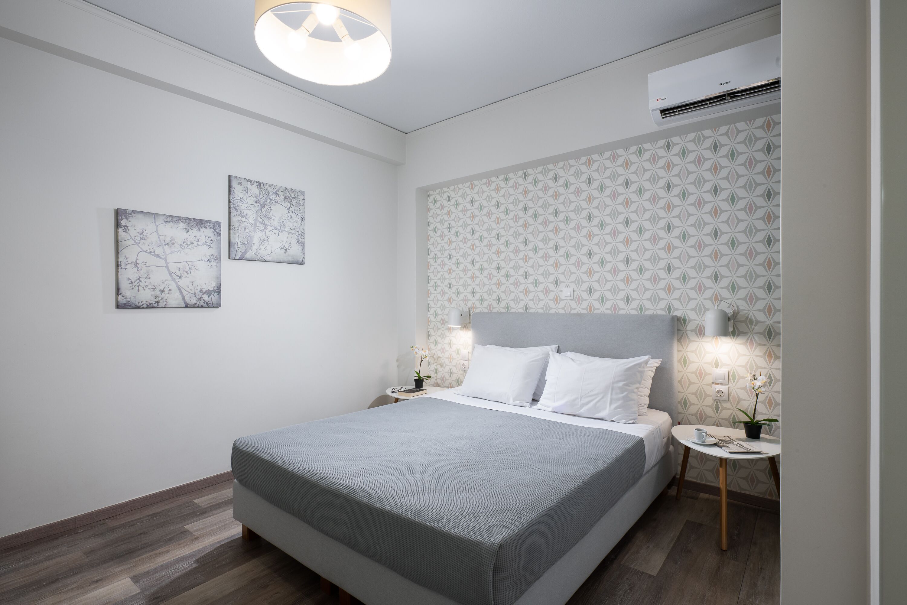 Ein modernes Schlafzimmer mit einem großen Bett, zwei an der Wand befestigten Kunstwerken, einer gemusterten Tapete, einem Nachttisch mit Lampe und einer Klimaanlage.